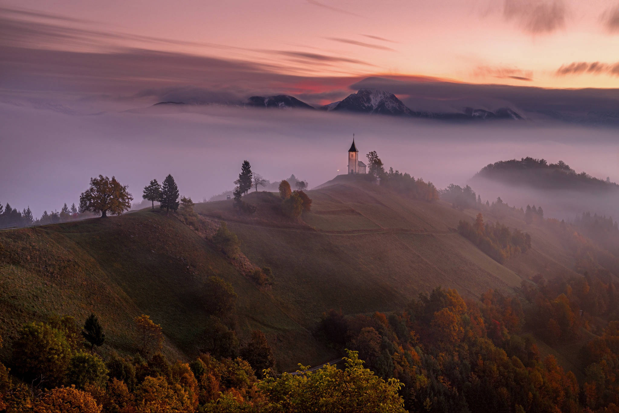 Slovenia 桌布 壁紙 — 黎明時分，一座孤獨的小教堂坐落在霧氣瀰漫的山丘頂上，秋樹和遠處的雪山在柔和粉紅和橙色光芒中閃耀。