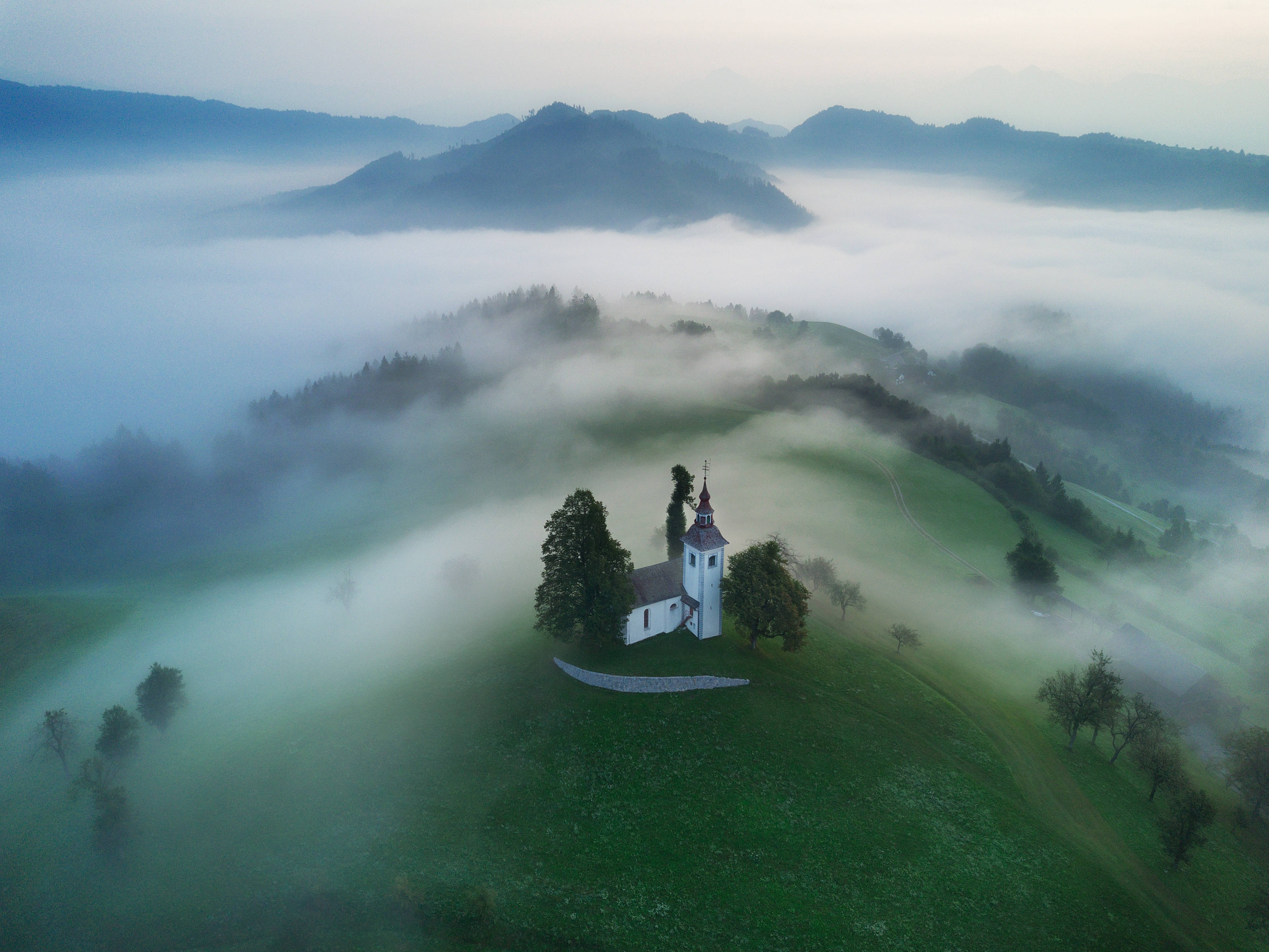 Slovenia 桌布 壁紙 — 黎明時分，一座白色小教堂坐落在霧氣瀰漫的綠色山丘上，周圍是雲霧繚繞的山巒的鳥瞰圖。