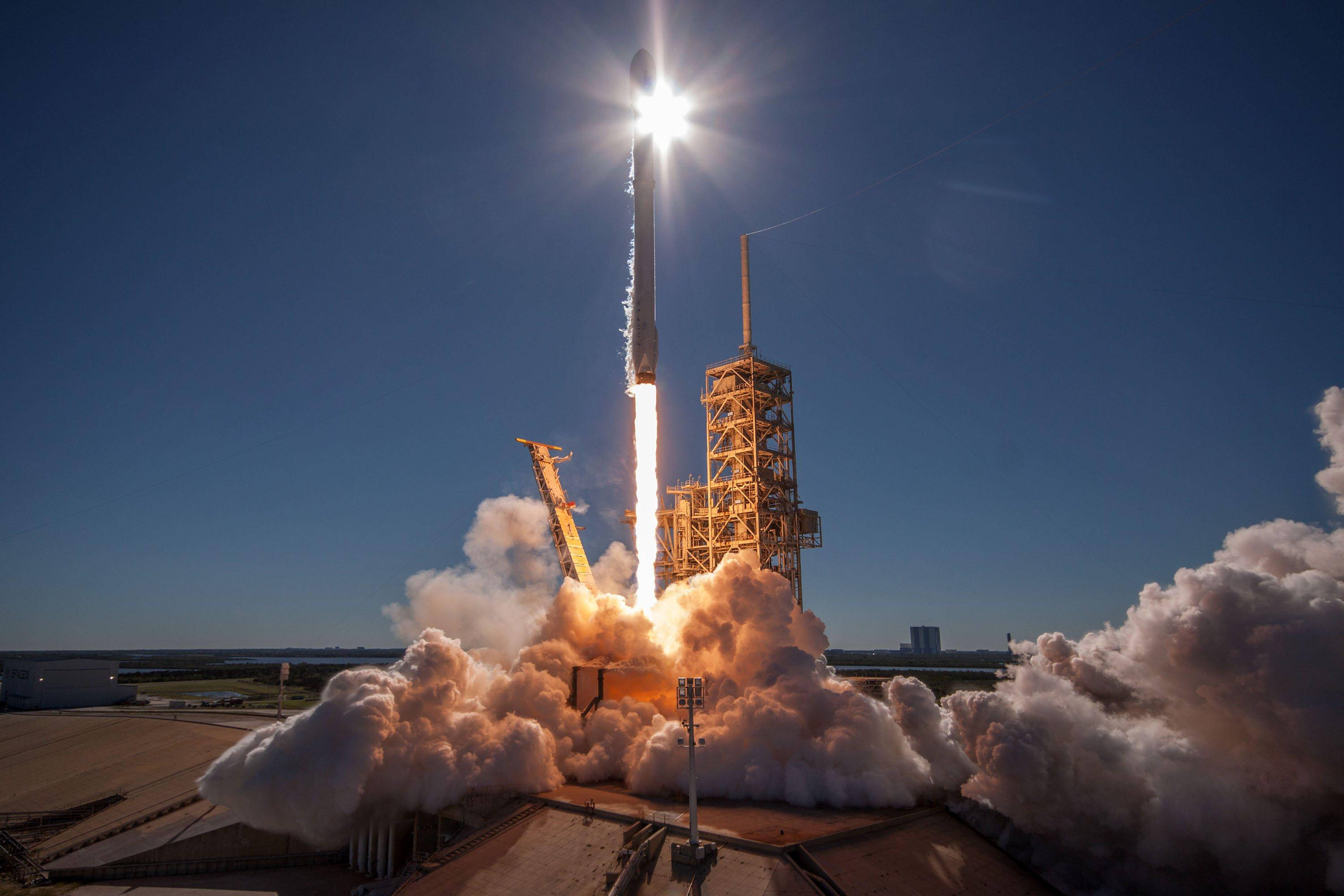 SpaceX 桌布 壁紙 — 黎明時分，一枚火箭在清澈的藍天中發射，伴隨著強烈的火焰和滾滾濃煙。