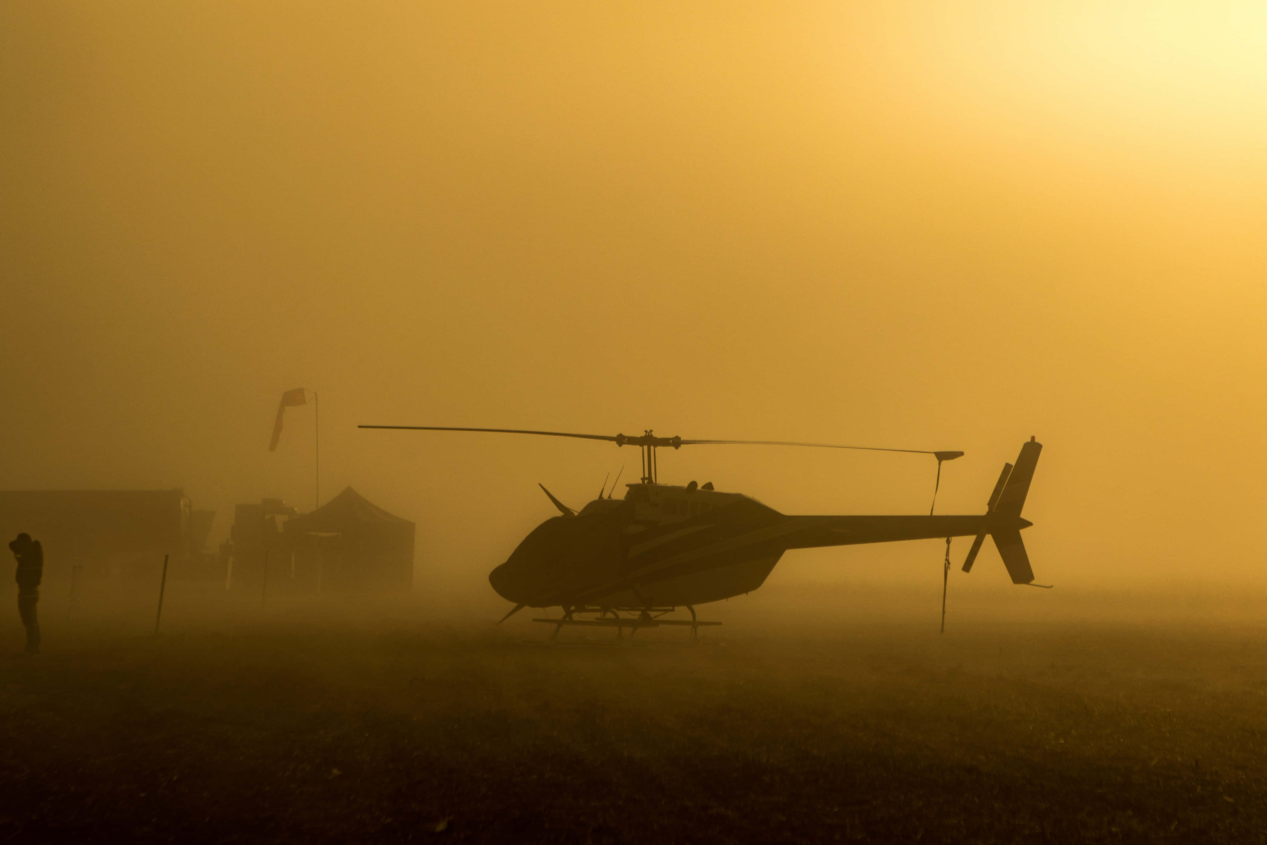 Helicopter 桌布 壁紙 — 黎明時分，一架直升機的剪影在濃厚的金色薄霧中靜止不動，附近有一個孤獨的身影在行走。