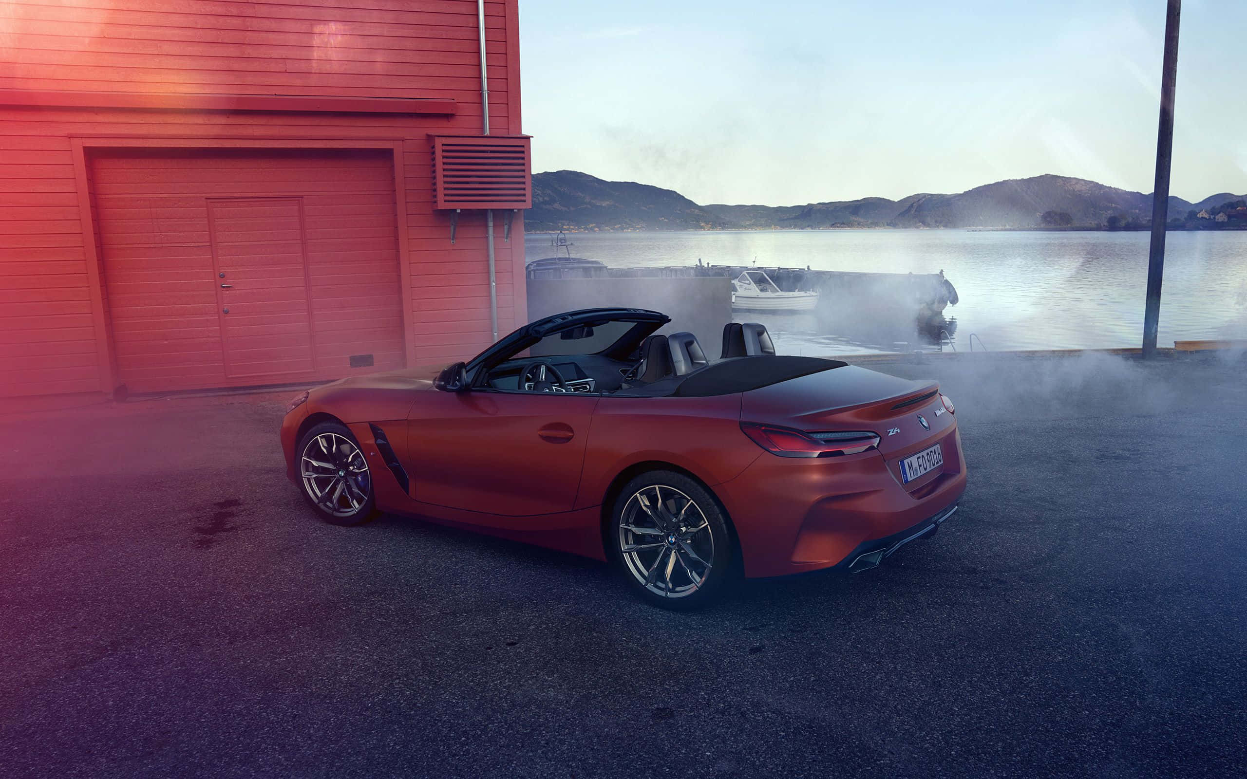 Bmw Z4 桌布 壁紙 — 黎明時分，一輛時尚的紅色敞篷跑車停放在紅色車庫旁，水面升起薄霧，背景中有船隻。