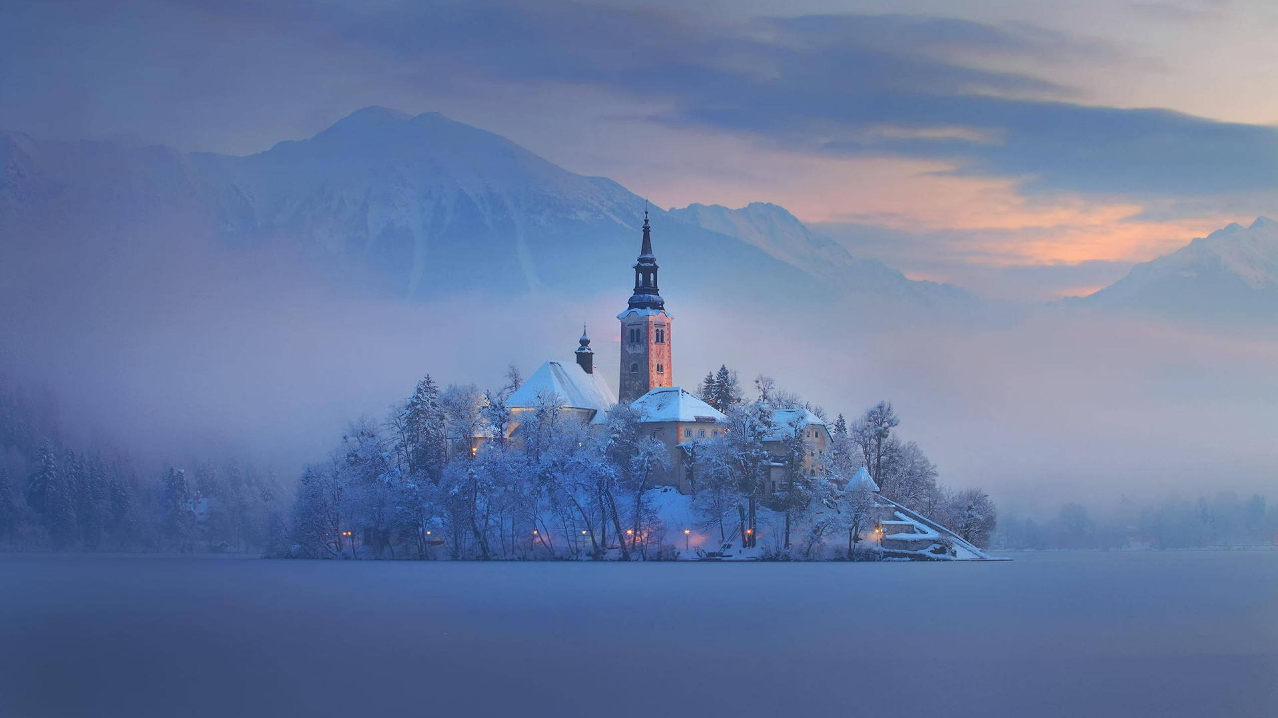 Slovenia 桌布 壁紙 — 黎明時分，冰凍湖面上的一座被雪覆蓋的教堂，背景是霧氣繚繞的山巒。