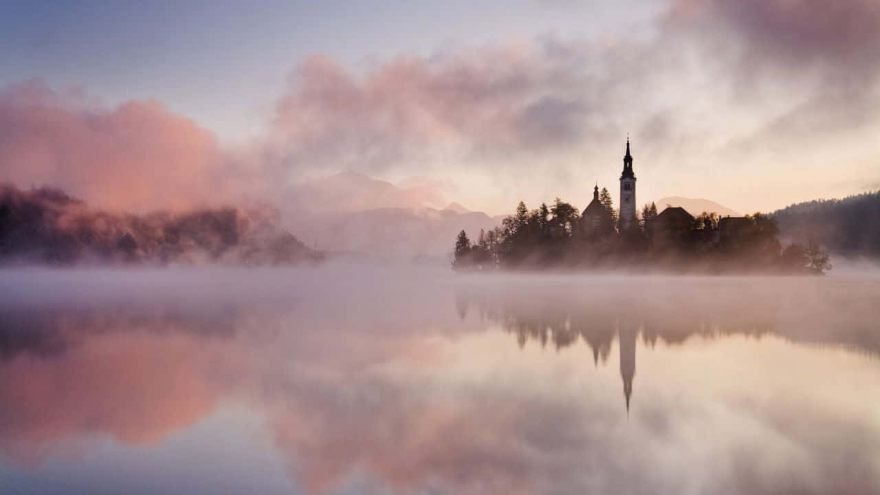 Slovenia 桌布 壁紙 — 黎明時分，寧靜的湖面上，教堂島嶼倒映在霧氣瀰漫的水中，天空呈現柔和的粉紅色和紫色。