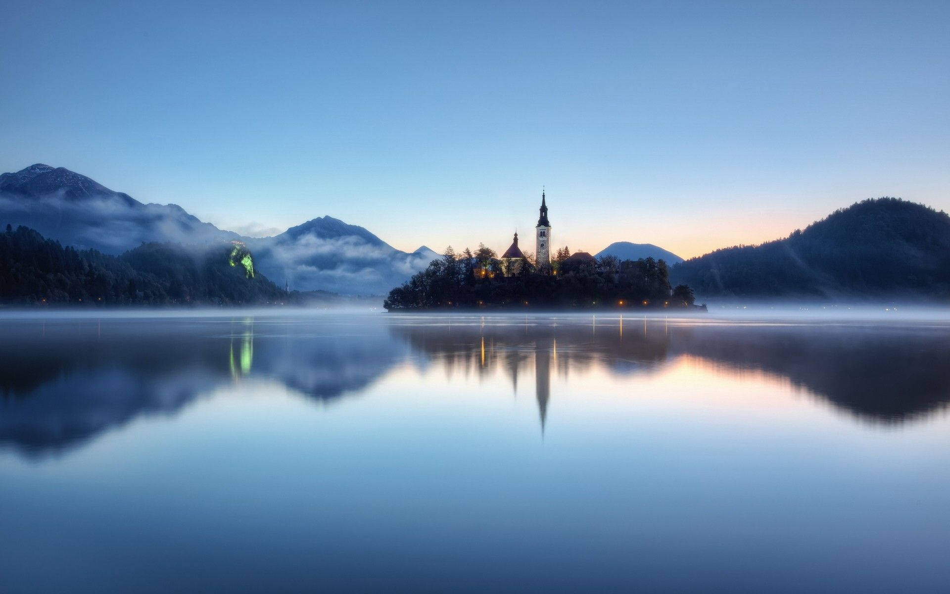 Slovenia 桌布 壁紙 — 黎明時分，寧靜的湖面倒映著霧氣籠罩的島嶼教堂，教堂燈光閃爍，背景是霧濛濛的山巒。