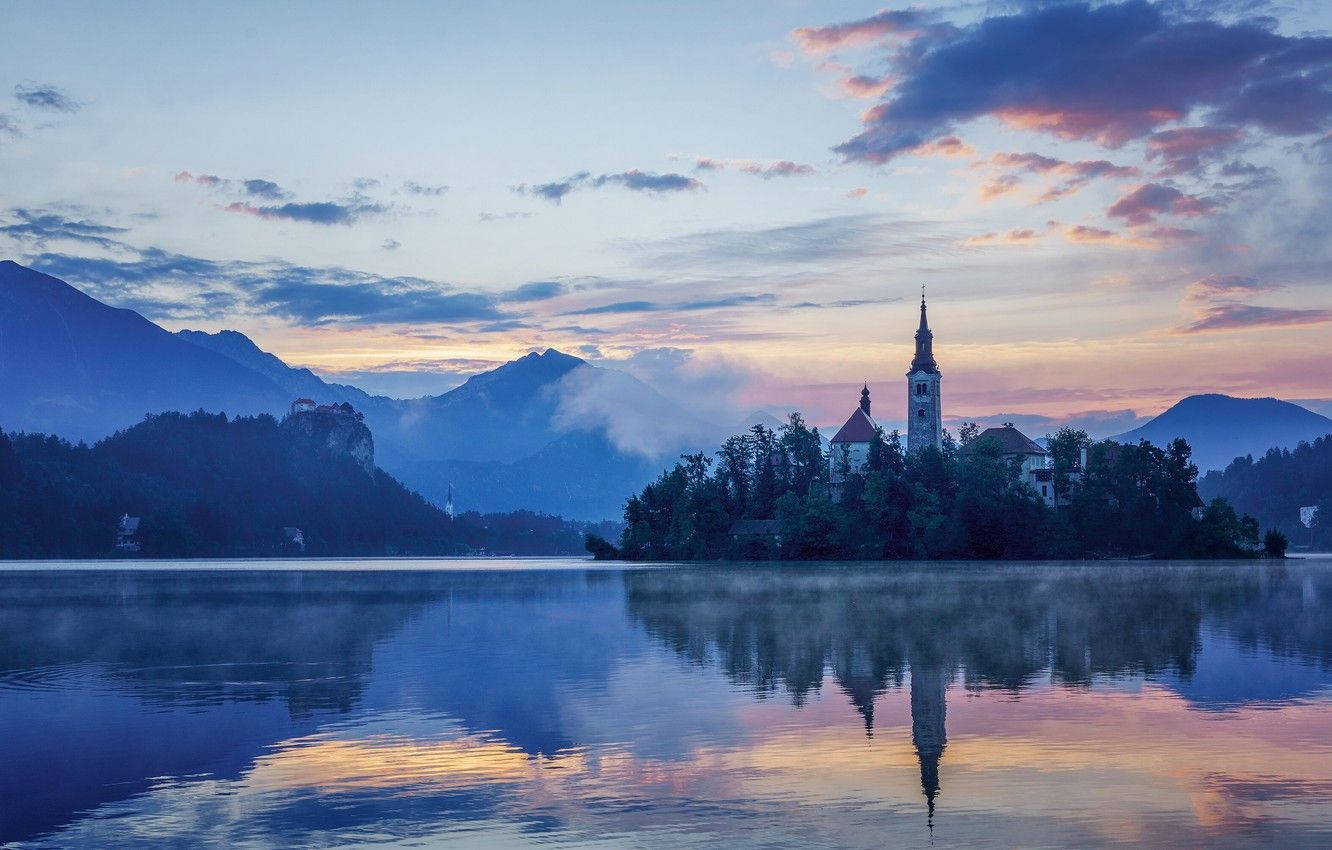 Slovenia 桌布 壁紙 — 黎明時分，寧靜的湖面映照著一座被霧氣繚繞的山巒環繞的島嶼上的教堂，天空呈現柔和色彩。