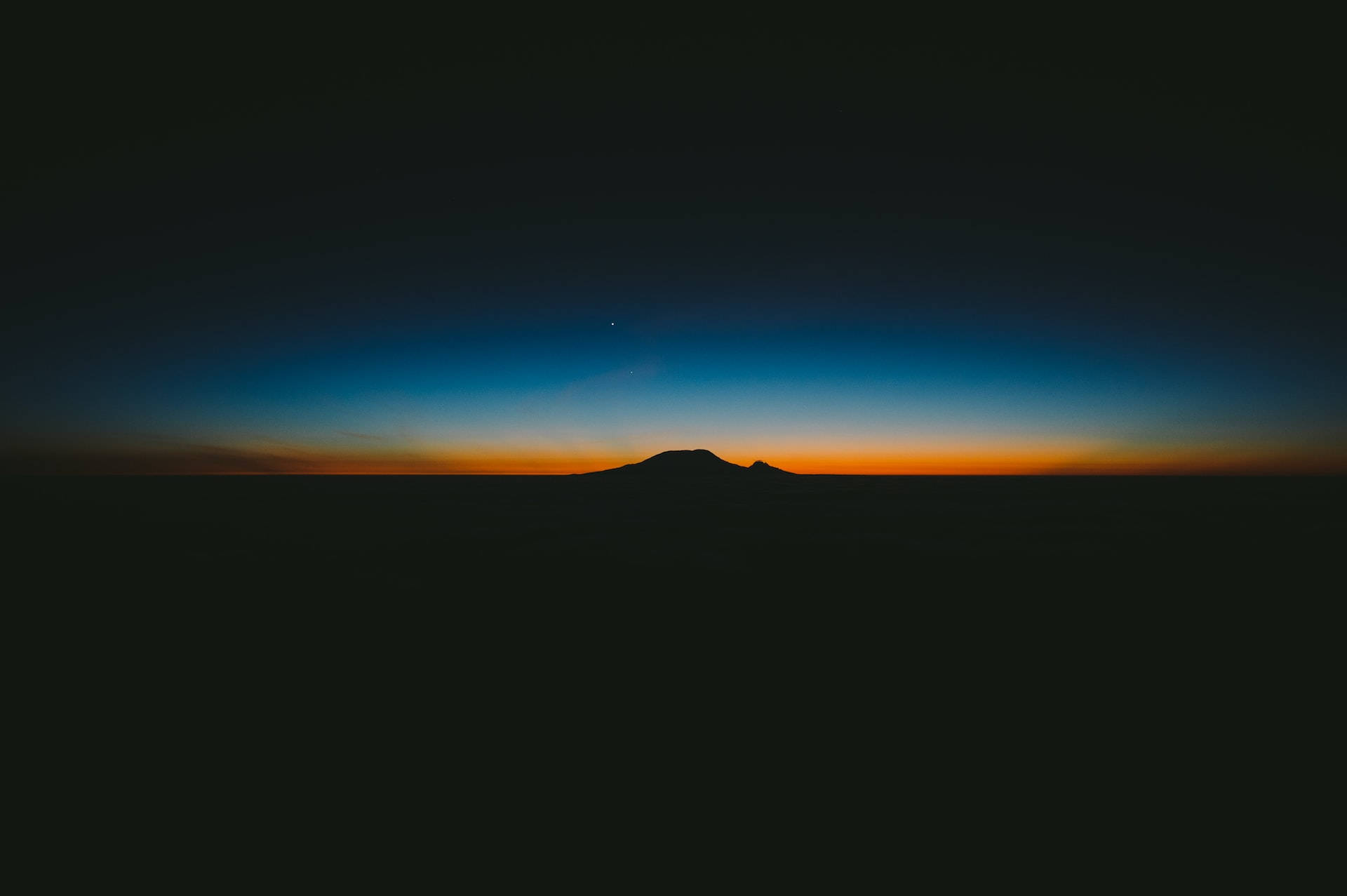 夜空 桌布 壁紙 — 黎明時分，山巒剪影映襯著從深藍漸變至火橙色的天空。