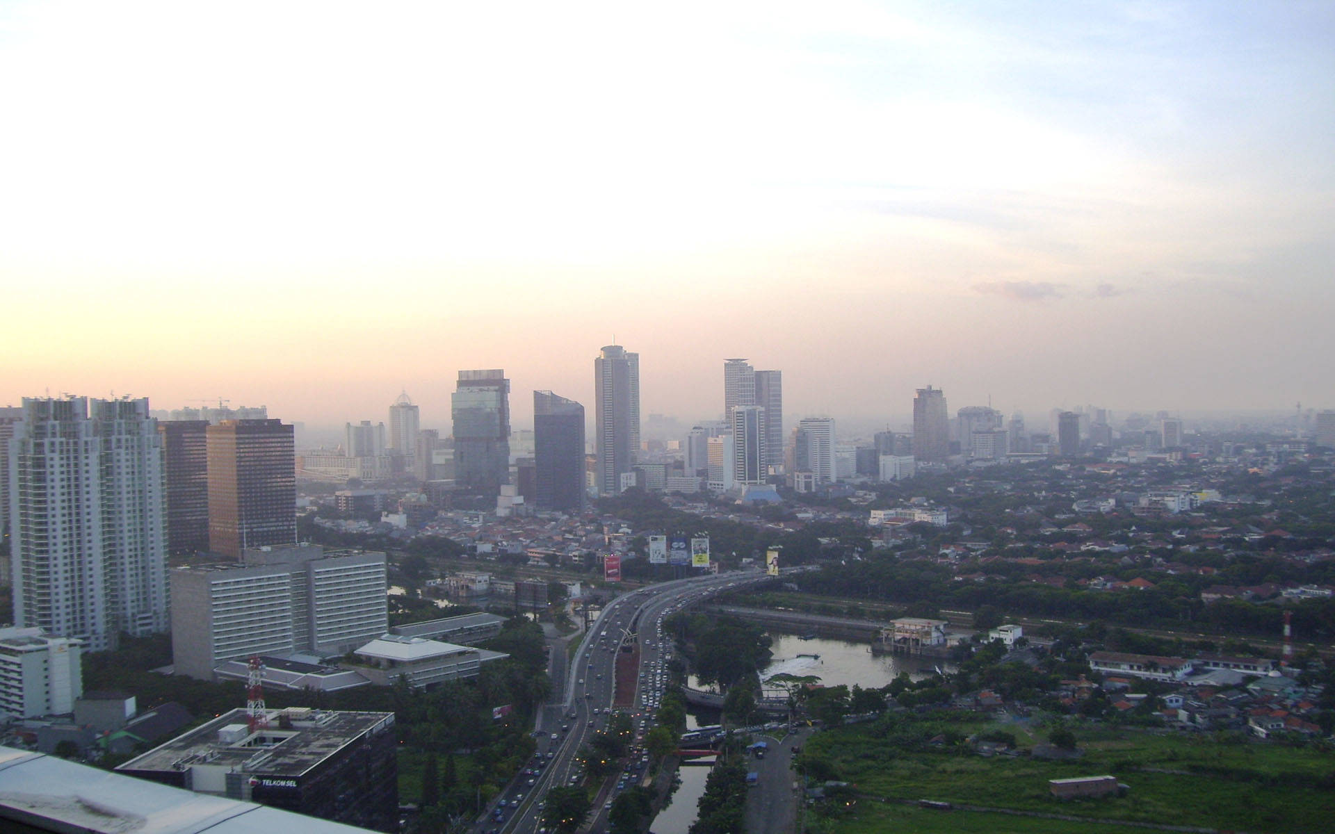 Jakarta 桌布 壁紙 — 黎明時分，廣闊的城市天際線，高聳的摩天大樓、繁忙的高速公路和蜿蜒穿過城市景觀的河流。