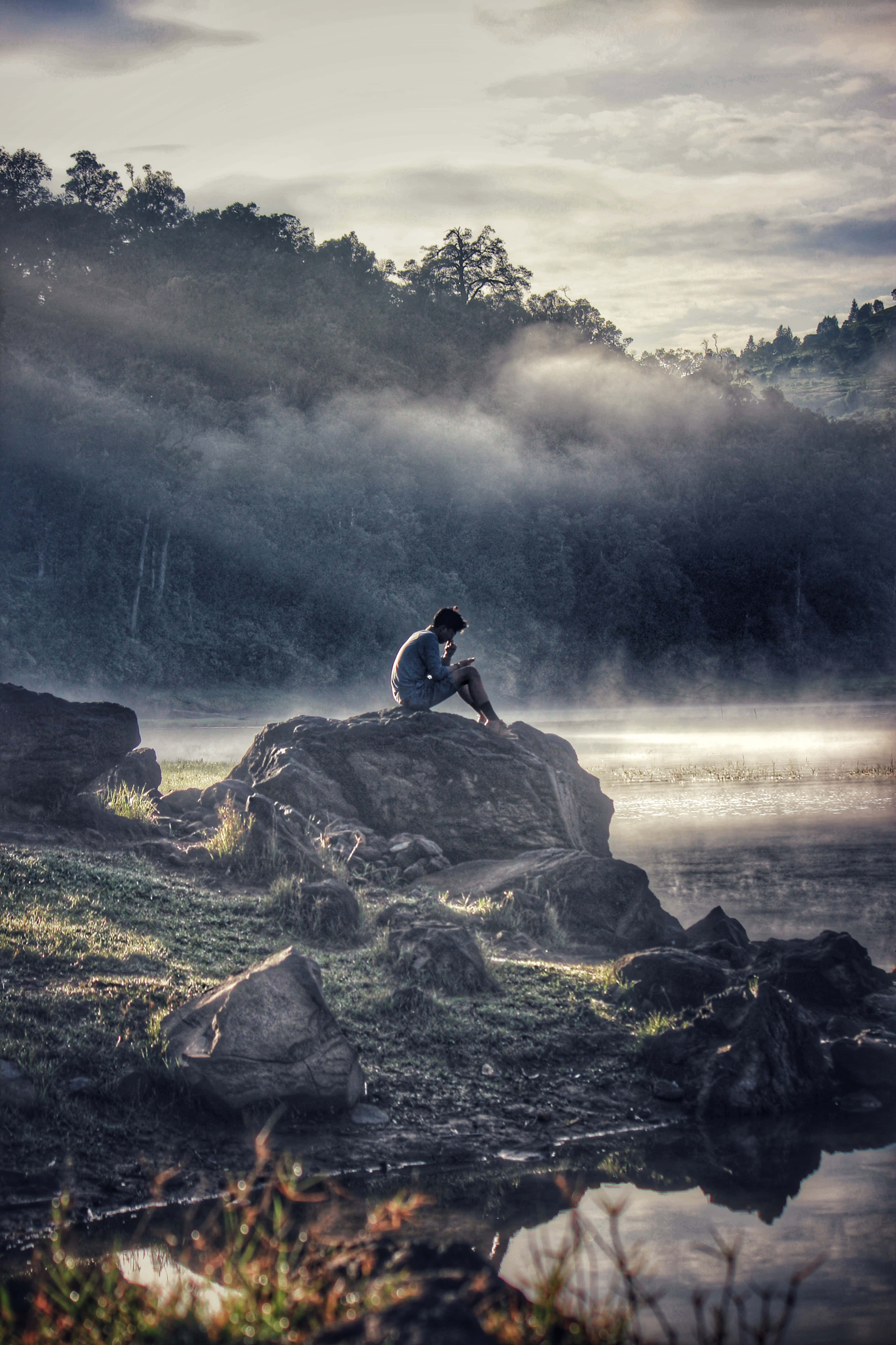 Sitting Alone 桌布 壁紙 — 黎明時分，薄霧籠罩著平靜的湖面，四周森林環繞，一個孤獨的身影坐在長滿青苔的石頭上沉思。