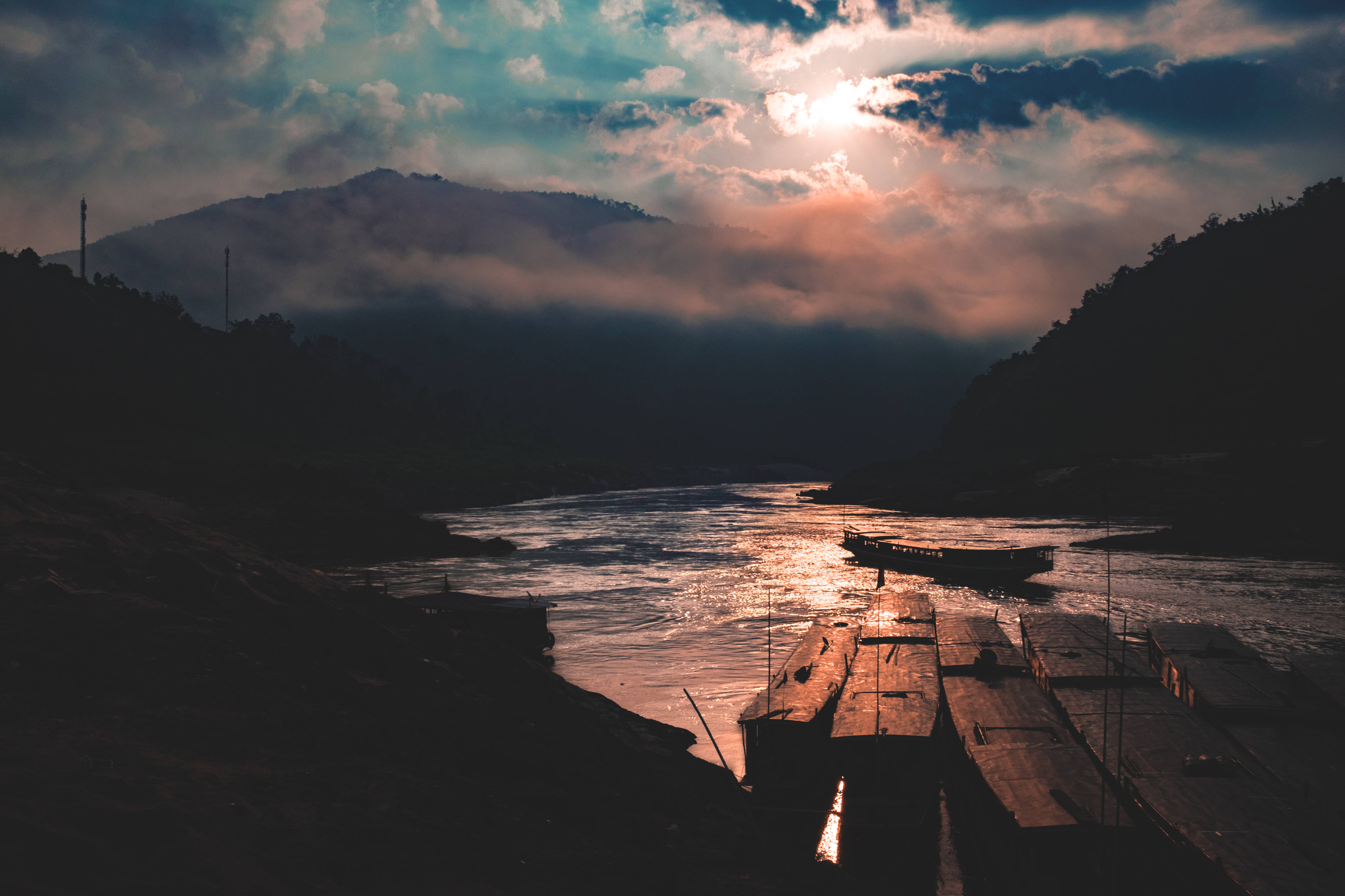 Laos 桌布 壁紙 — 黎明時分，陽光穿透雲層，在黑暗森林覆蓋的山丘之間蜿蜒的河流上投下金色光芒。