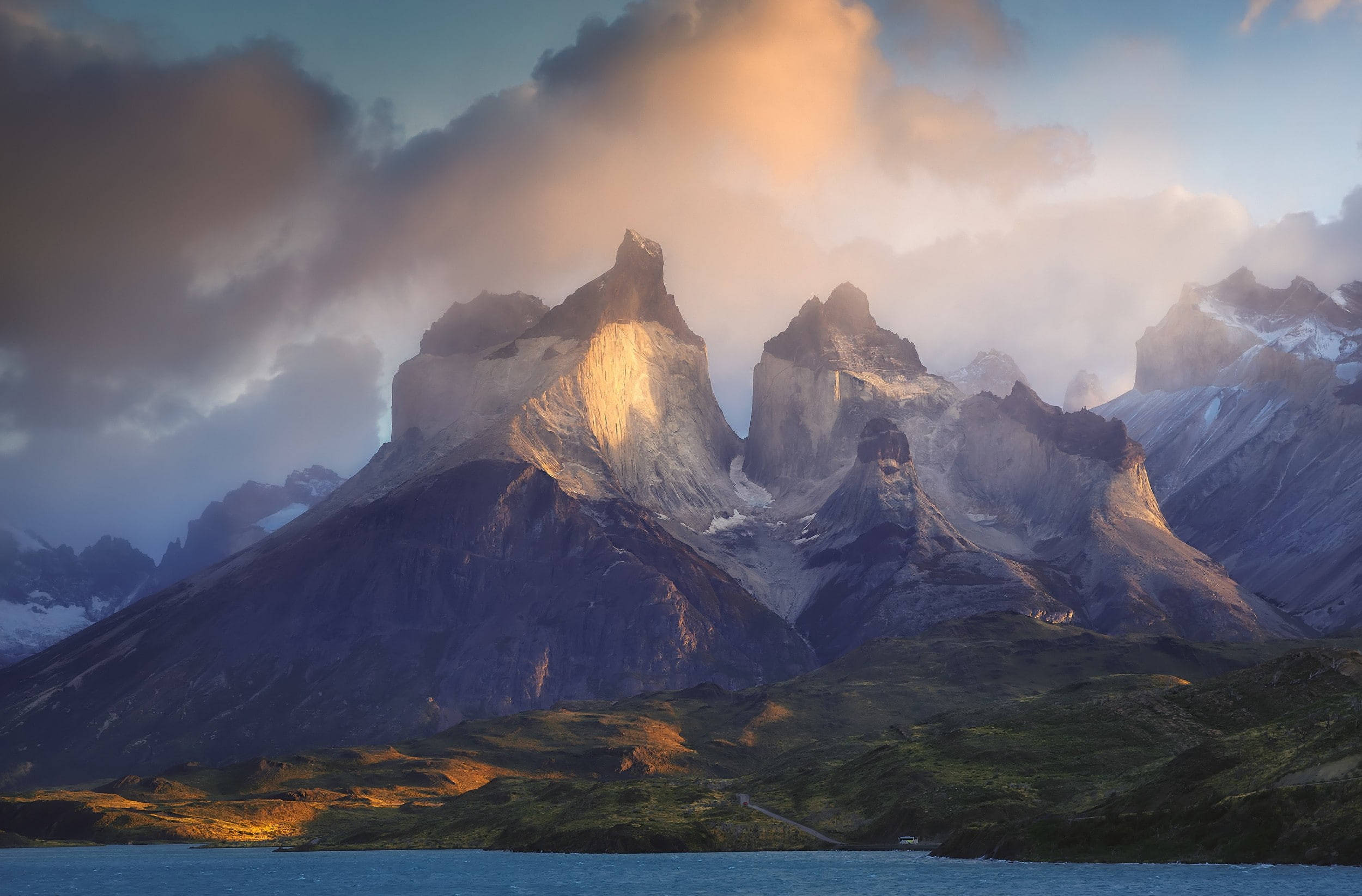 Patagonia 桌布 壁紙 — 黎明時分，陽光穿過雲層，照亮了平靜的藍色湖泊上方崎嶇的山峰。