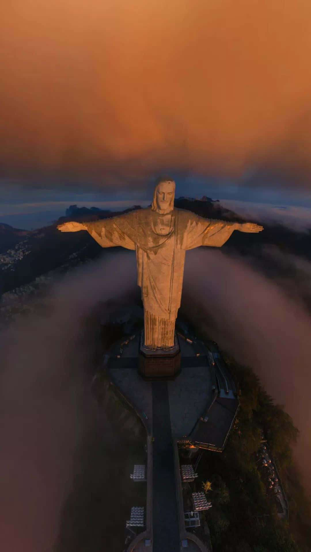 Jesus iPhone 桌布 壁紙 — 黎明時分基督像的鳥瞰圖，金色光芒照亮其雙臂，背景是霧氣繚繞的山巒和橙色的天空。