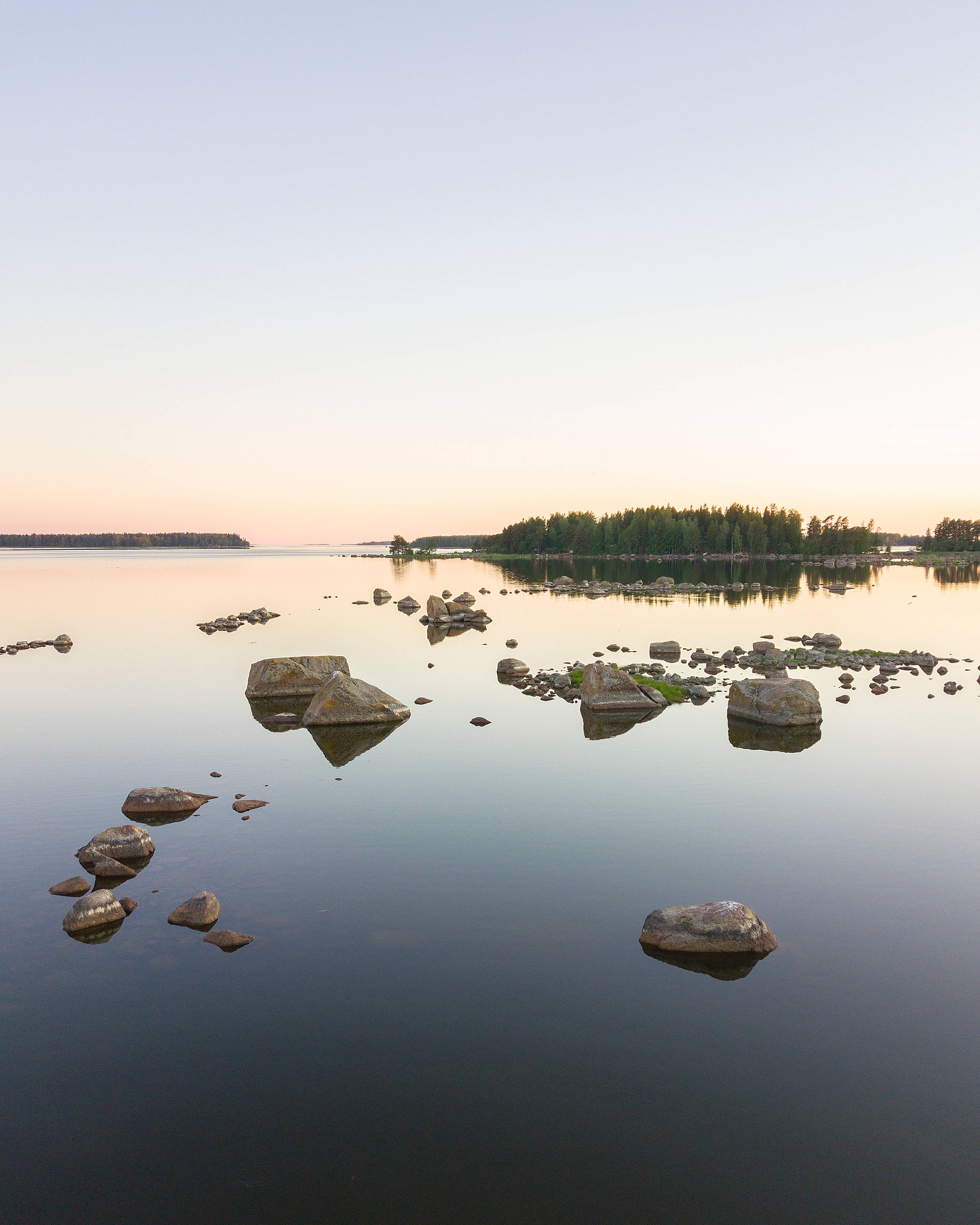 Finland 桌布 壁紙 — 黎明時分的寧靜湖泊，散落的岩石在平靜的水面上倒映，遠處樹木成蔭的湖岸，柔和粉彩的天空。