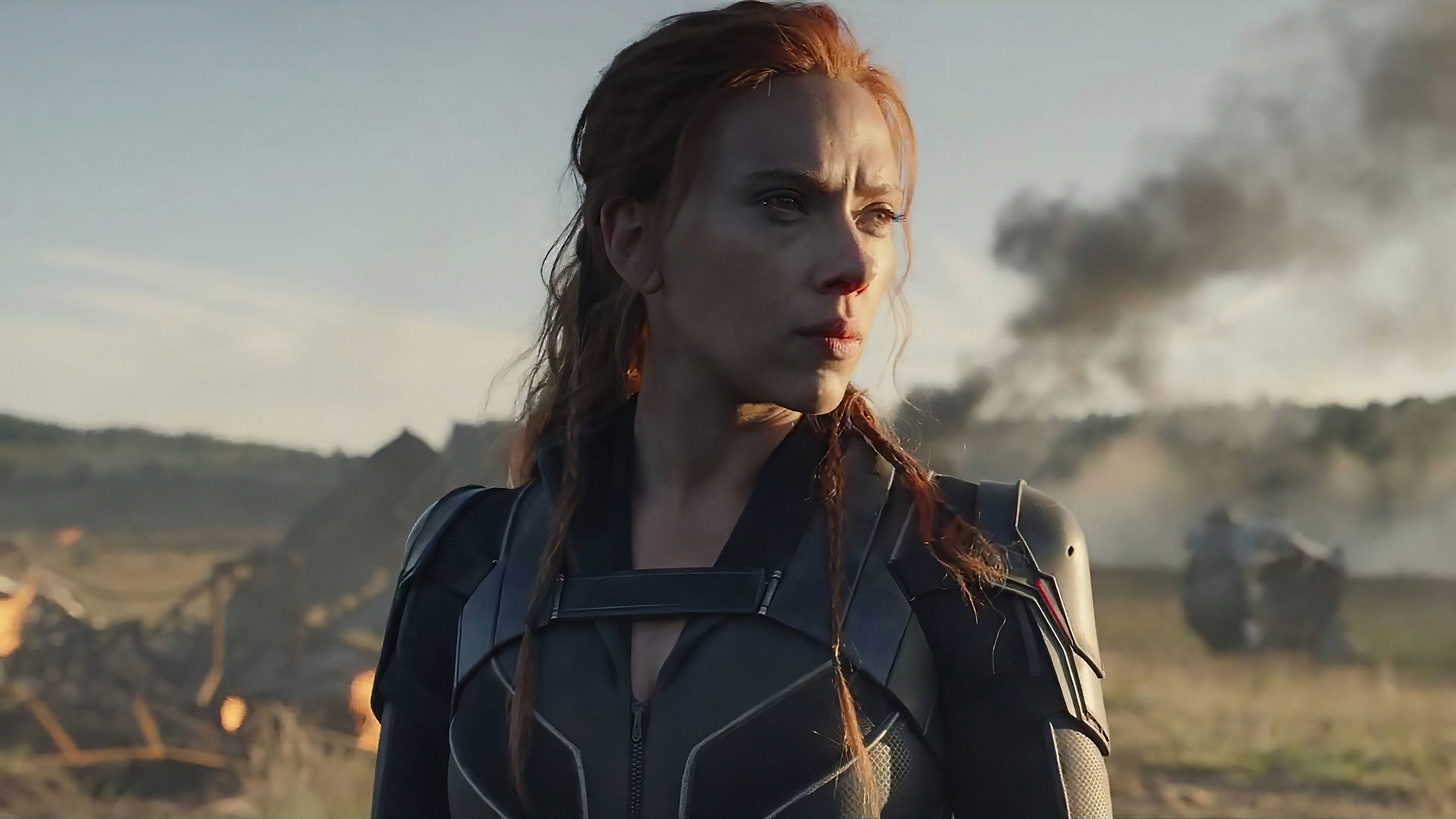 Scarlett Johansson Black Widow 桌布 壁紙 — 黑寡婦站在一個被戰爭摧毀的田野中，身後煙霧升騰，她專注地凝視前方。
