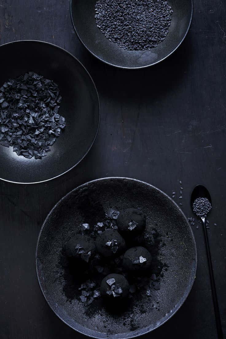 Black Garlic 桌布 壁紙 — 黑巧克力松露搭配黑芝麻和炭片，擺放在霧面黑碗中，置於有紋理的表面上。