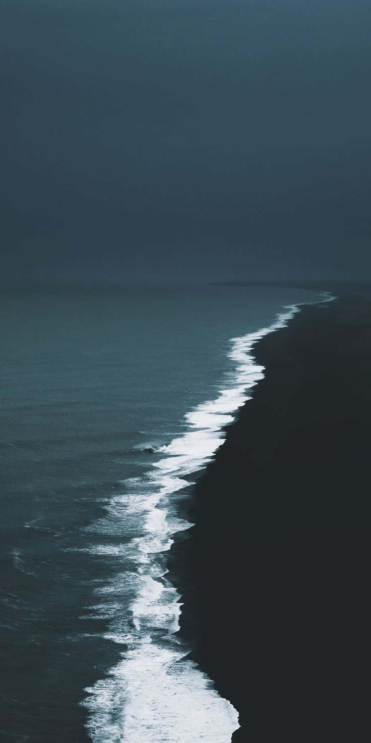 Dark Ocean 桌布 壁紙 — 黑暗的暴風雨海洋沖刷著黑色火山沙灘，陰沉天空下白色泡沫勾勒出海岸線。