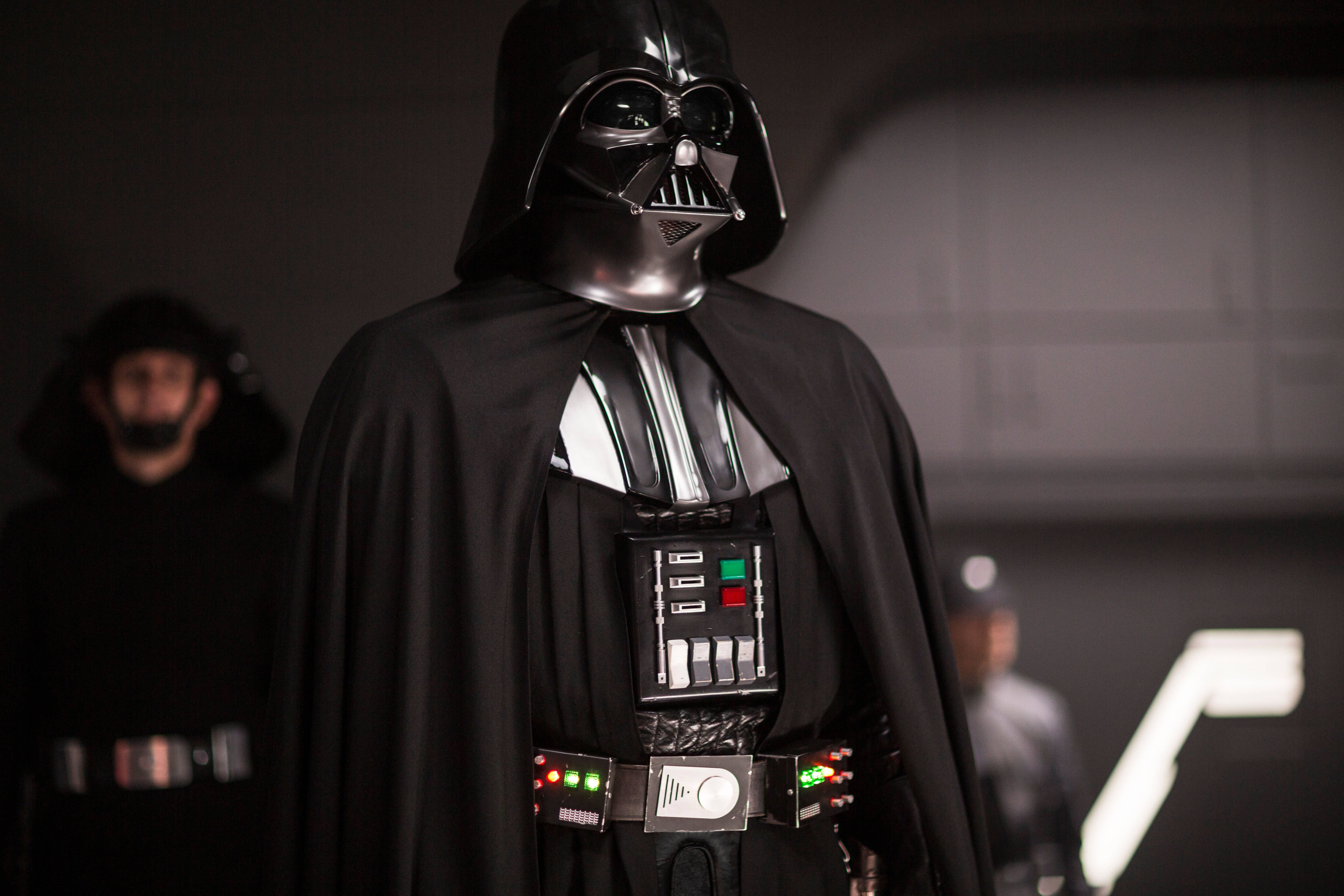 Vader 桌布 壁紙 — 黑武士達斯維達身穿全副黑色盔甲和披風，威嚴地站在燈光昏暗的科幻走廊中，身後有模糊的風暴兵。