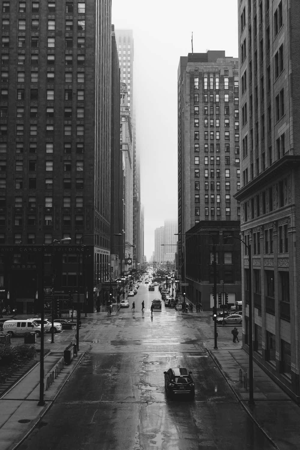 Black And White Street 桌布 壁紙 — 黑白城市街景，由高聳建築物框住，濕漉漉的路面反射城市燈光及稀疏的行人。