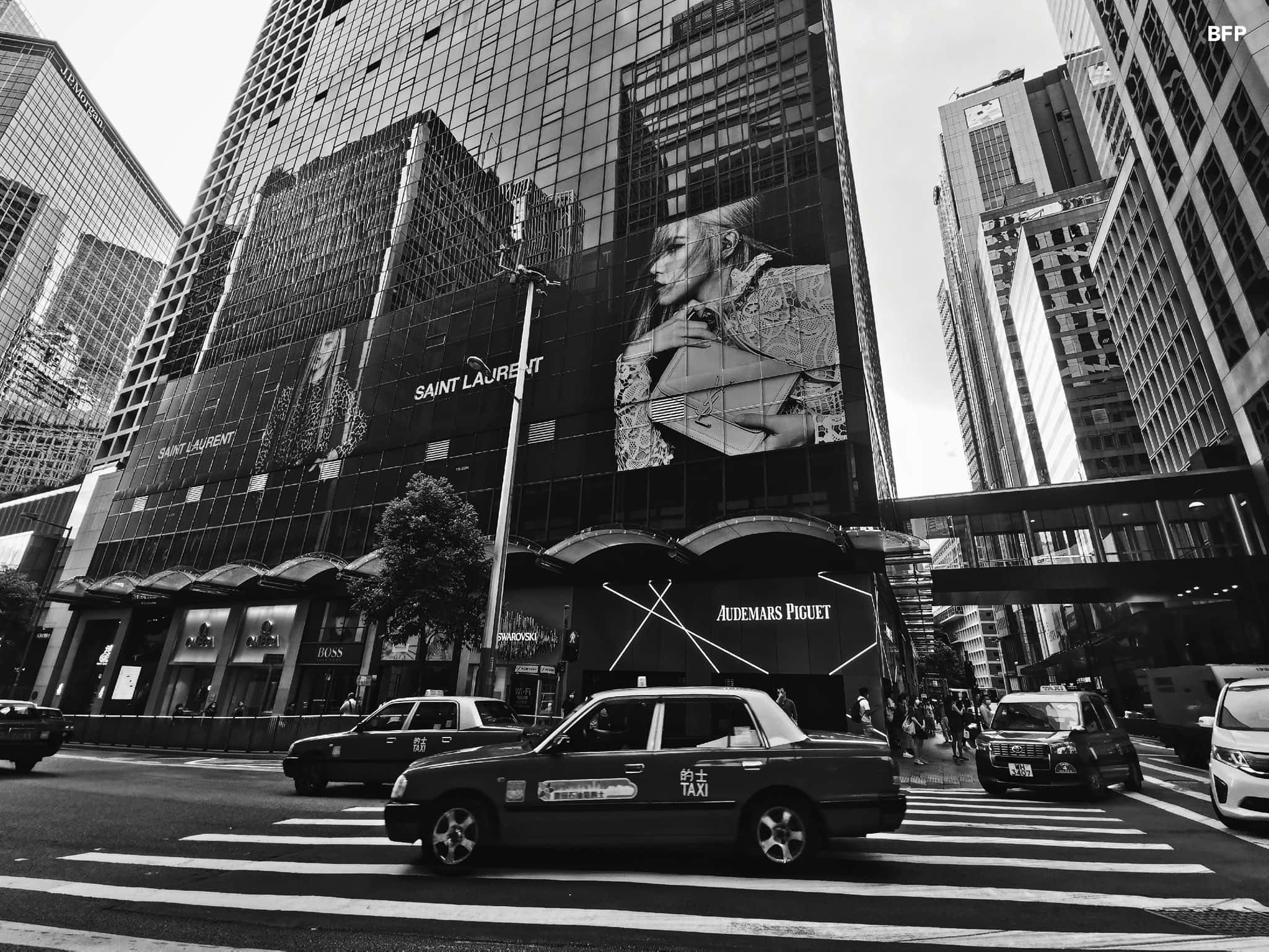 YSL 桌布 壁紙 — 黑白城市街景，計程車在玻璃摩天大樓下穿過斑馬線，大樓上掛滿奢侈品牌廣告牌。