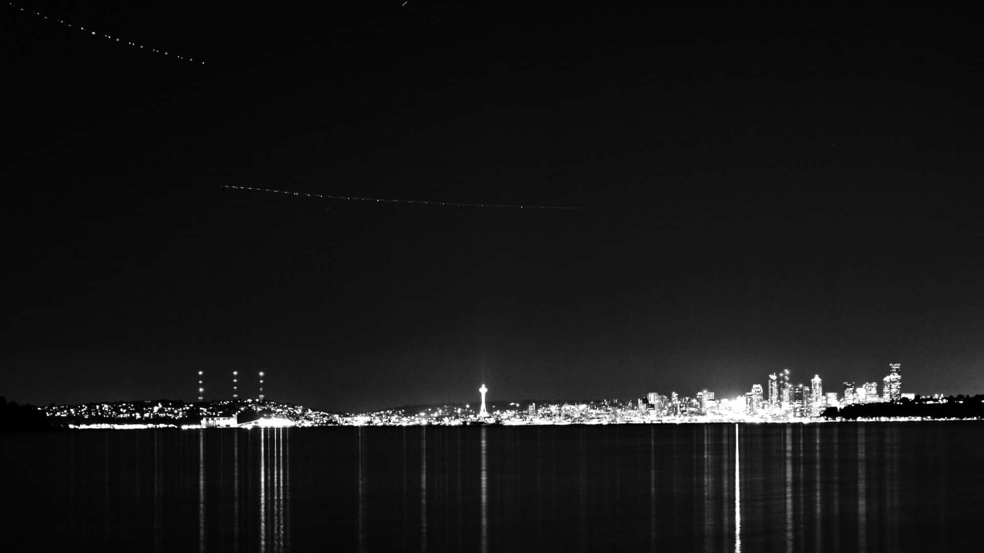 Seattle Black And White 桌布 壁紙 — 黑白夜景，城市天際線倒映在平靜的水面上，飛機尾跡劃過漆黑的天空。