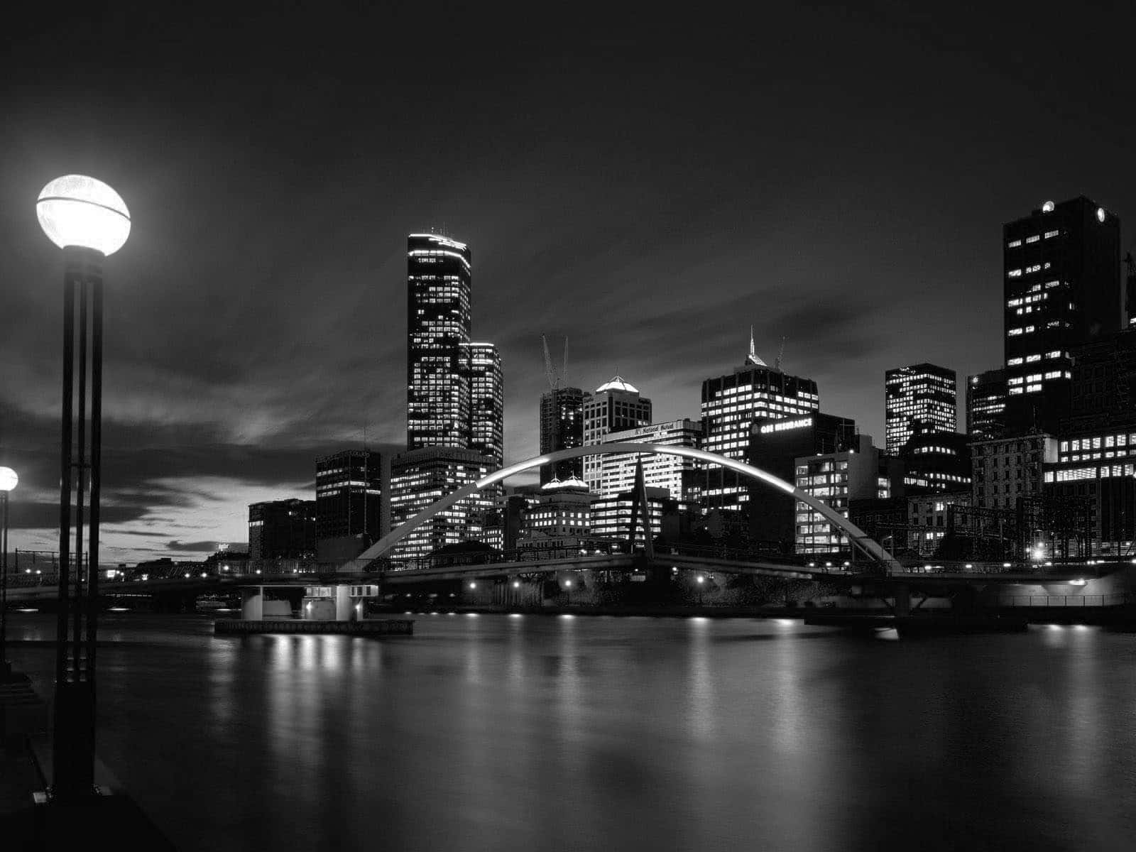 Black And White Cityscape 桌布 壁紙 — 黑白夜景，雲天下水面倒映摩天大樓，一座發光的橋樑橫跨水上。