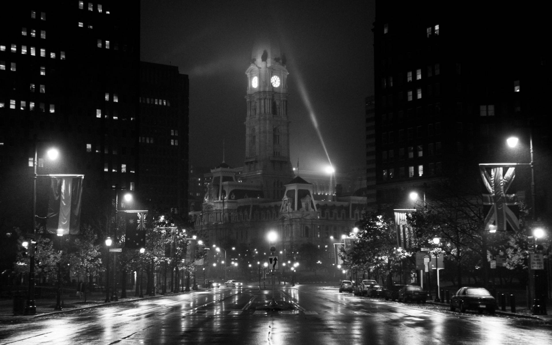 Philadelphia 桌布 壁紙 — 黑白夜景城市景觀，特色是一個宏偉的鐘樓被聚光燈照亮，下方是一條濕漉漉、有倒影的街道，兩旁排列著建築物和街燈。