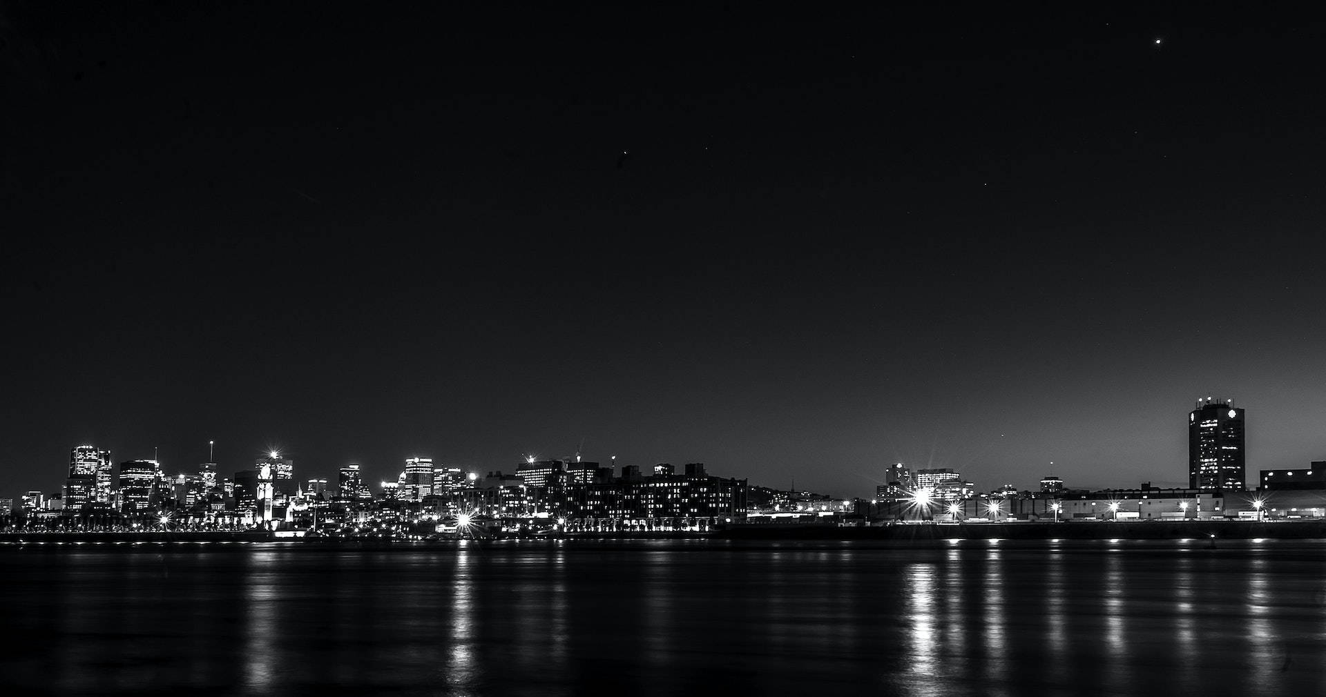 黑色 桌布 壁紙 — 黑白夜景城市景觀，發光的摩天大樓在星空下平靜的水面上倒映。