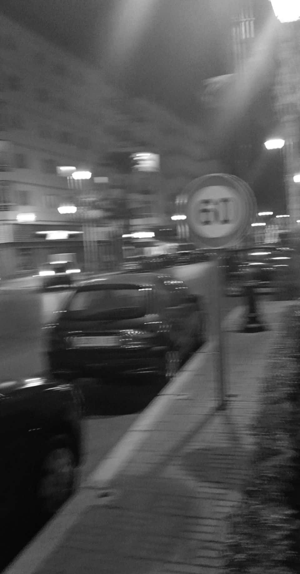 Aesthetic Black And White 桌布 壁紙 — 黑白夜間街景，帶有動態模糊的汽車和一個在街燈下發光的50速度限制標誌。