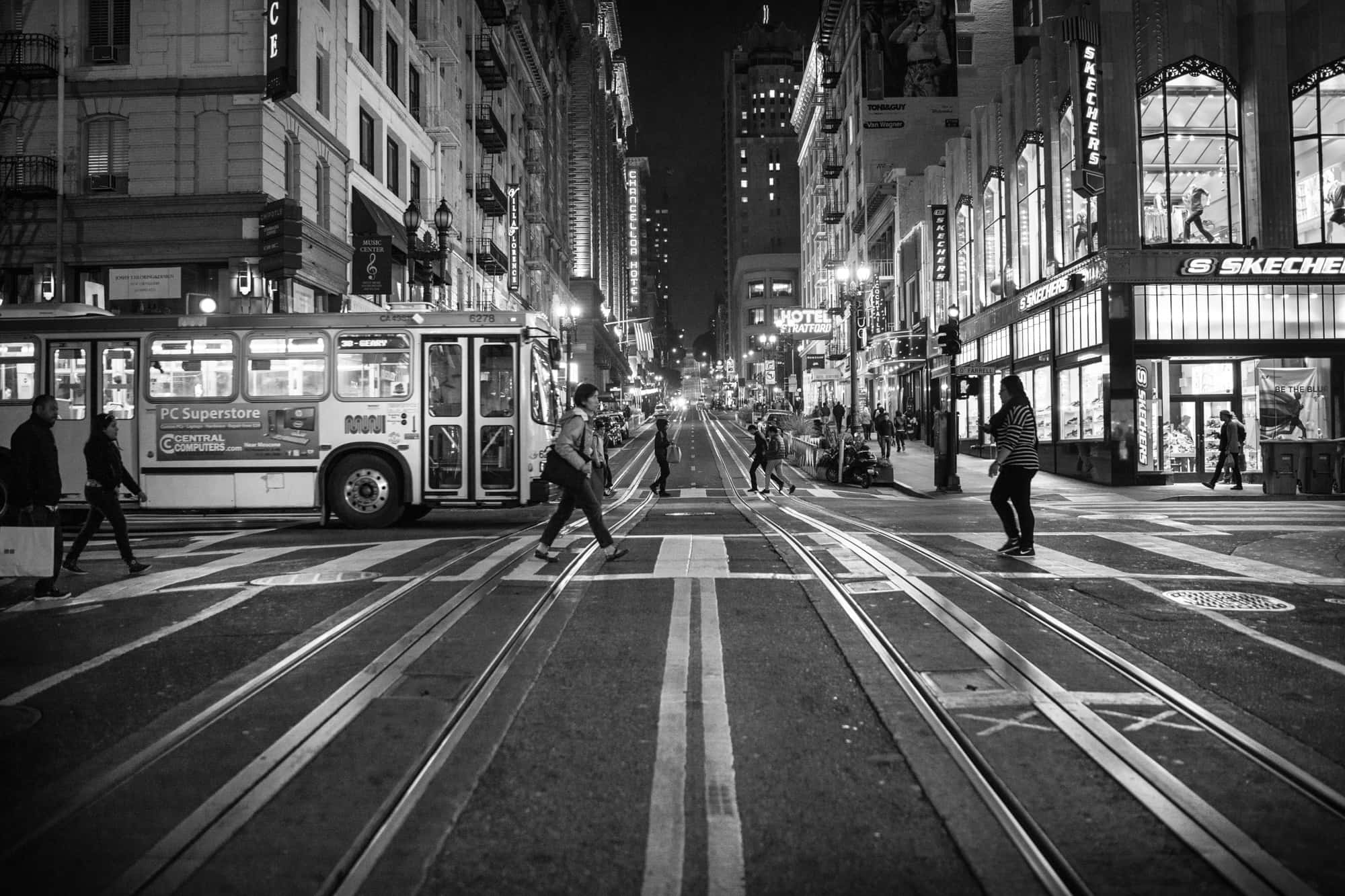 San Francisco Black And White 桌布 壁紙 — 黑白夜間街景，行人橫越軌道，無軌電車在繁華的城市峽谷中行駛。
