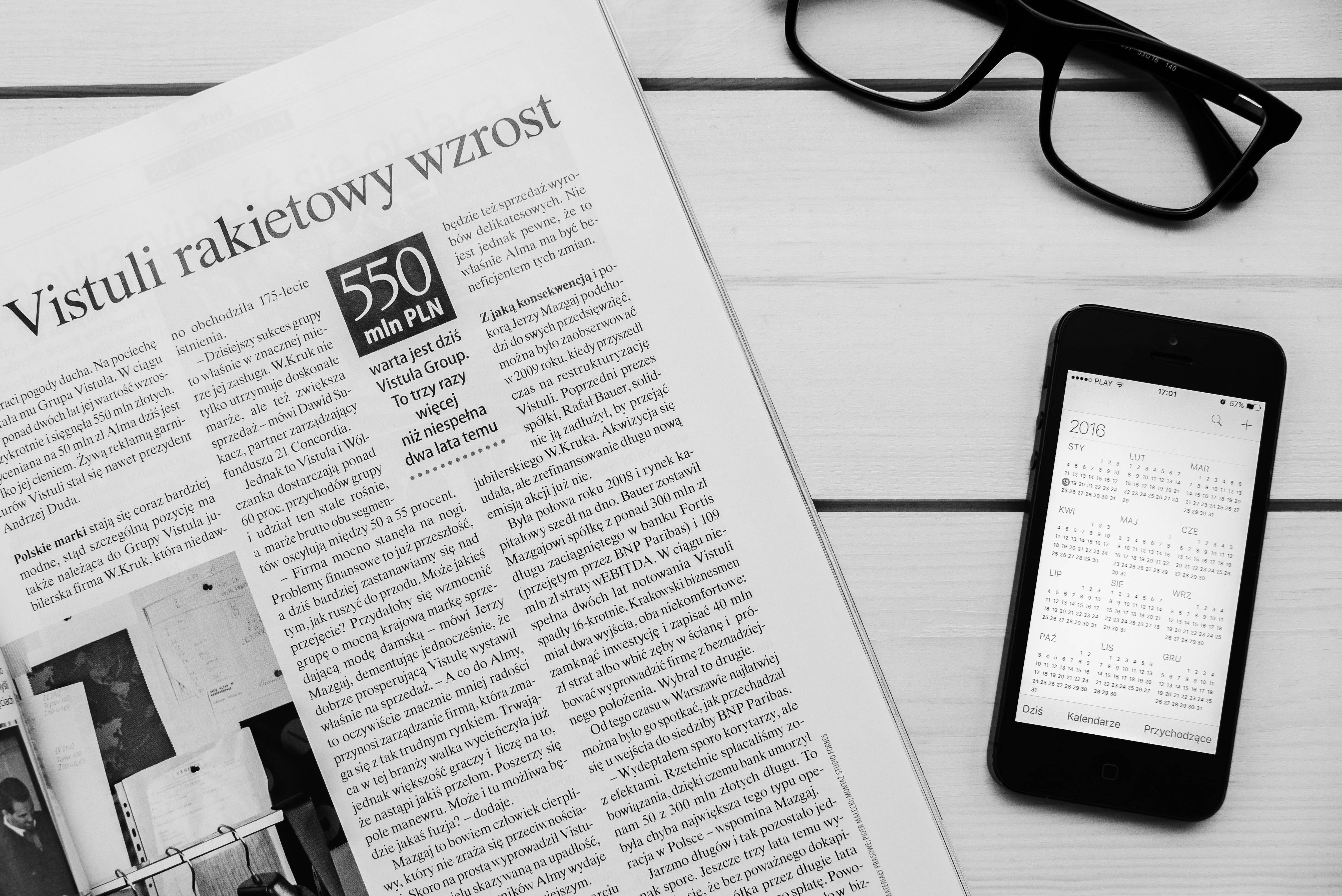 Newspaper 桌布 壁紙 — 黑白平鋪構圖：報紙文章、眼鏡及顯示日曆的智慧型手機置於木質表面上。