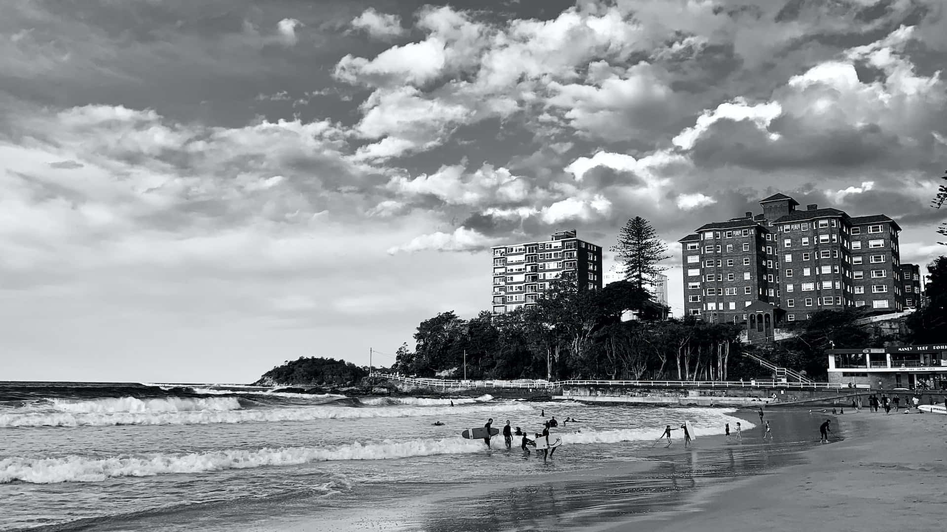 Black And White Beach 桌布 壁紙 — 黑白海岸場景，衝浪者在海浪中，建築物矗立在懸崖上，天空佈滿戲劇性的雲層。