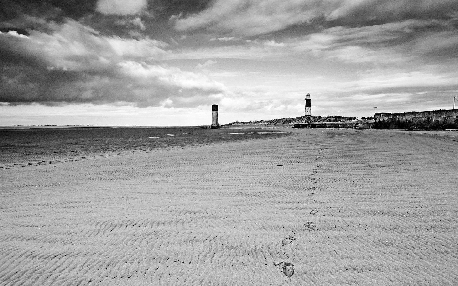Beach Desktop 桌布 壁紙 — 黑白海灘場景，腳印在戲劇性的多雲天空下通往兩座燈塔。