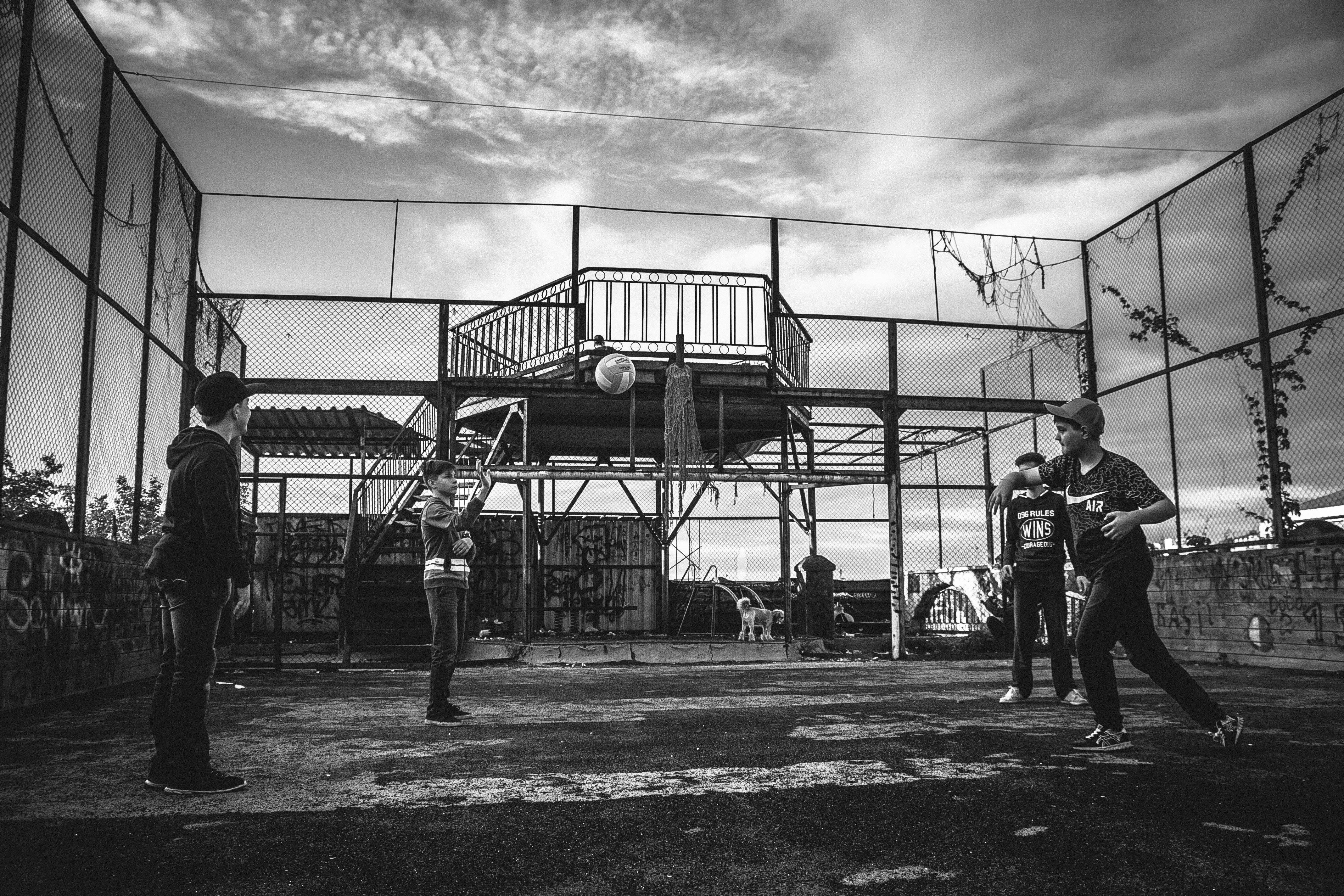 Playground 桌布 壁紙 — 黑白照片，四名年輕男子在多雲天空下，於一個圍欄圍著的粗糙城市球場打排球。