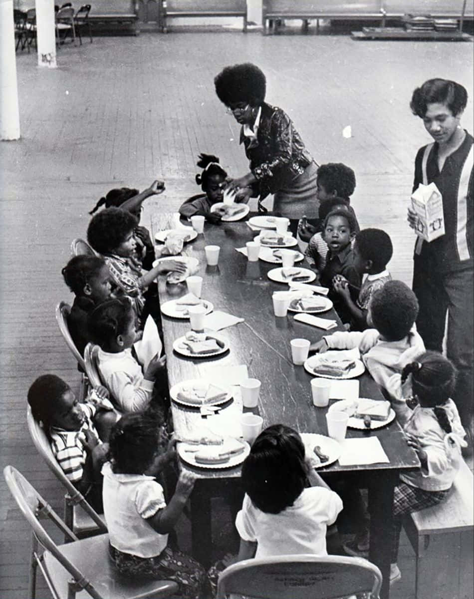 Black Panther Party 桌布 壁紙 — 黑白照片，孩子們坐在長桌旁吃午餐，有成年人在食堂為他們服務。