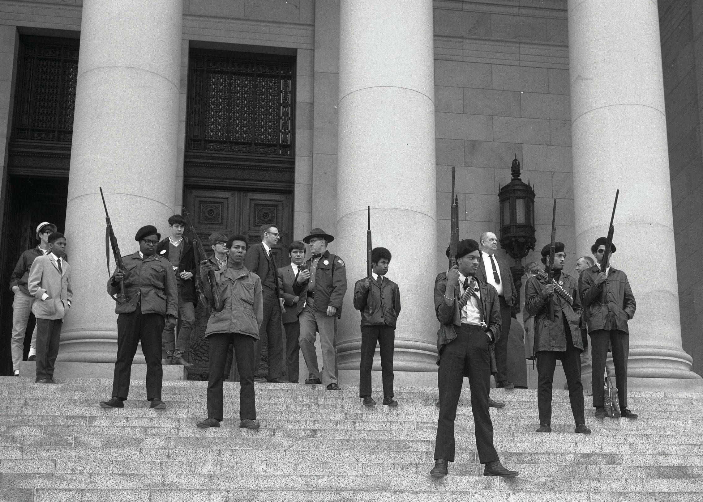Black Panther Party 桌布 壁紙 — 黑白照片，年輕男子身穿外套，手持步槍，站在宏偉柱廊前的石階上。