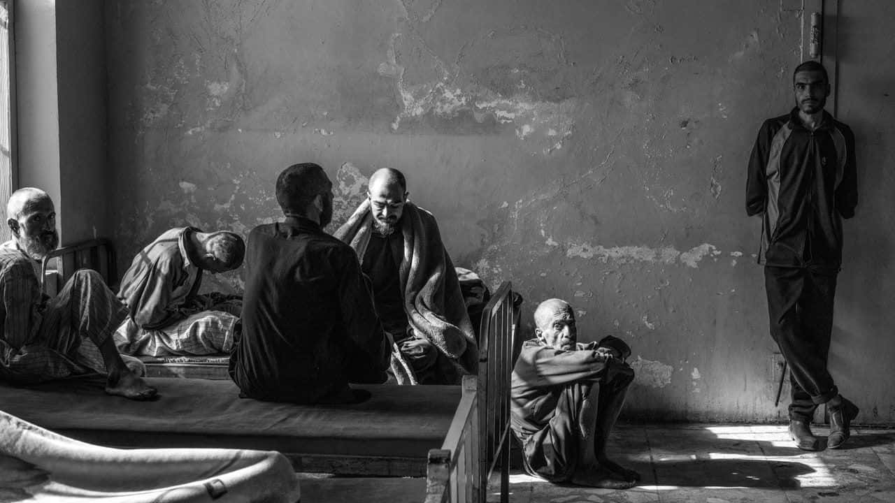 Taliban 桌布 壁紙 — 黑白照片，拍攝幾位長者在一間牆壁剝落、陽光斜照的破舊房間內休息和坐著。