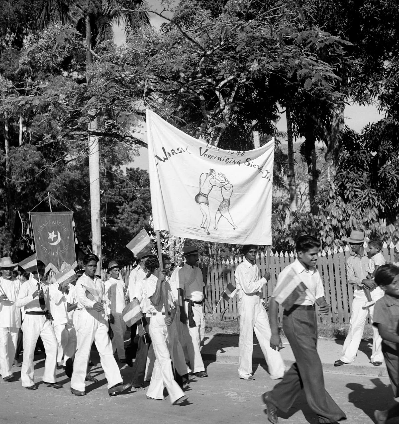1940s 桌布 壁紙 — 黑白照片，身穿白色制服的男人們手持旗幟和橫幅，沿著綠樹成蔭的街道遊行。