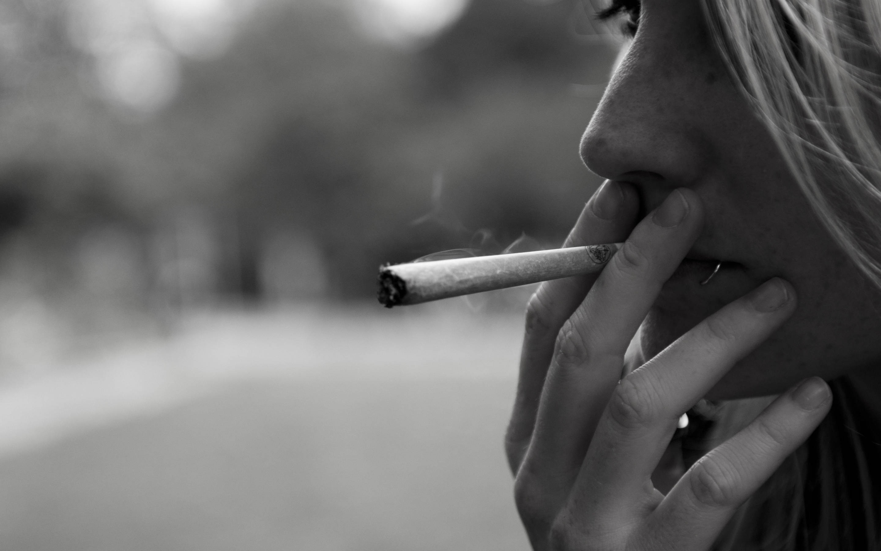 吸大麻 桌布 壁紙 — 黑白特寫，一個人抽煙，煙霧從煙頭升起，背景模糊的戶外環境。