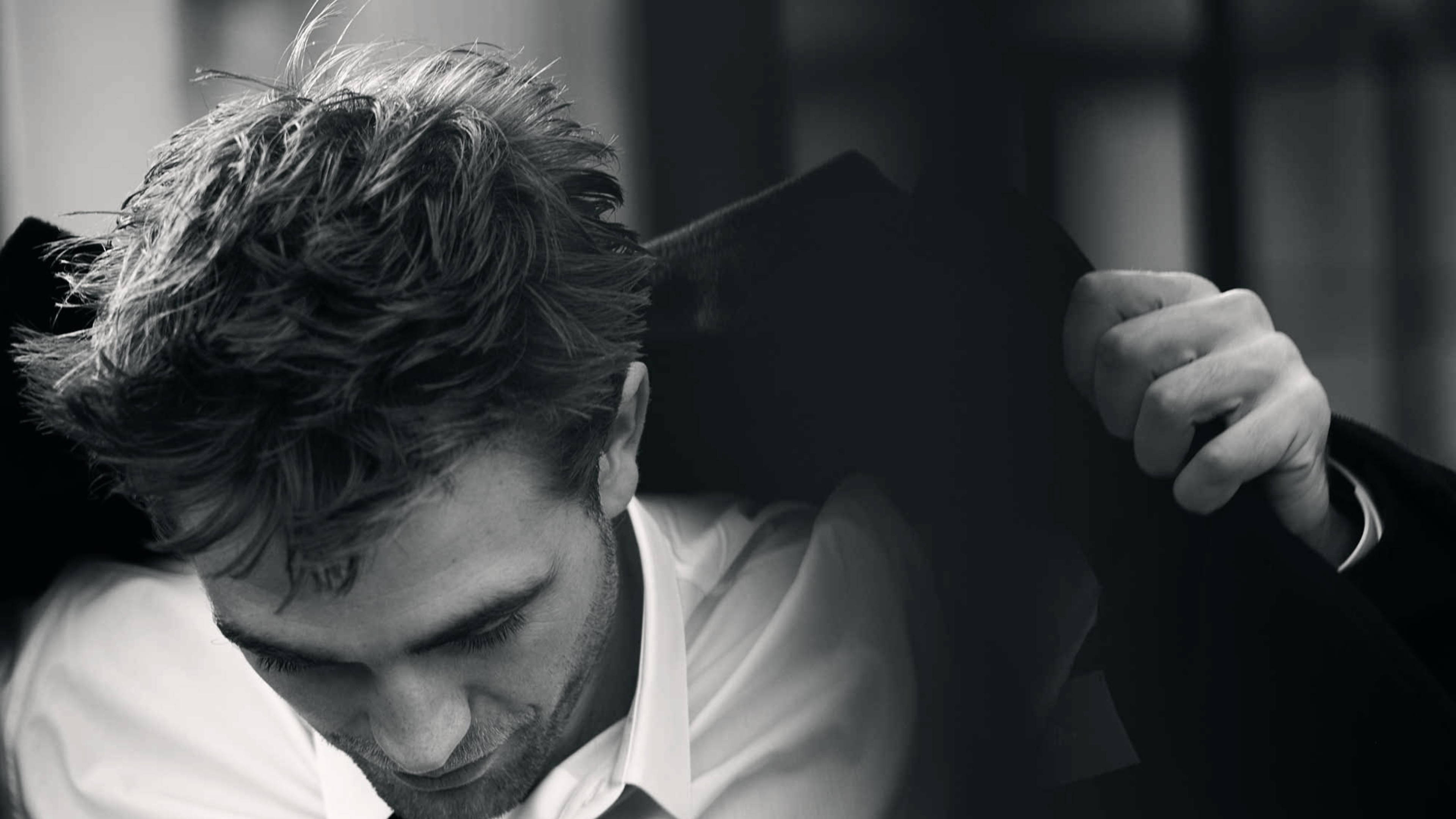 Robert Pattinson 桌布 壁紙 — 黑白特寫，一名頭髮凌亂的男子低頭，將深色外套搭在肩上。