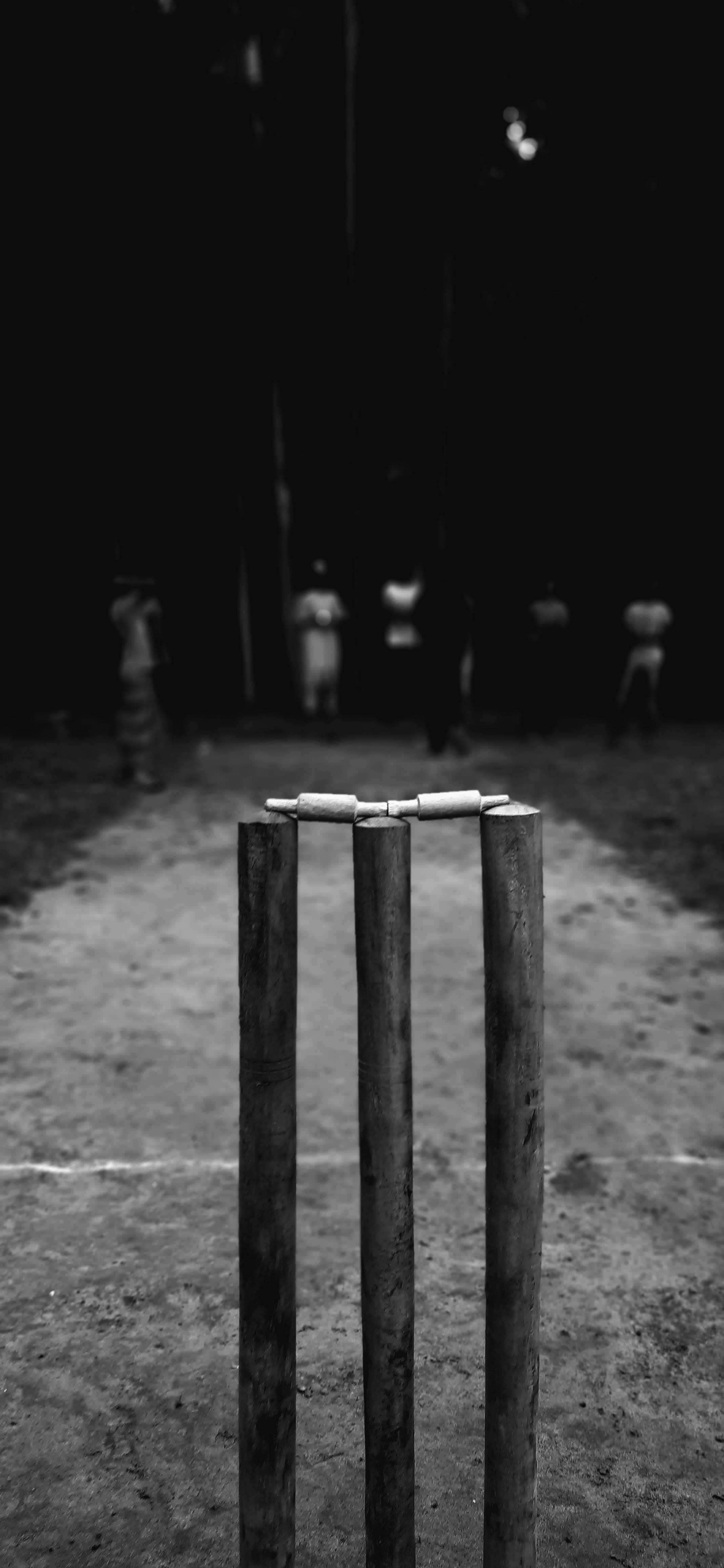 crickets 桌布 壁紙 — 黑白特寫，板球三柱門上放著橫木，背景是昏暗的戶外場地和模糊的球員。