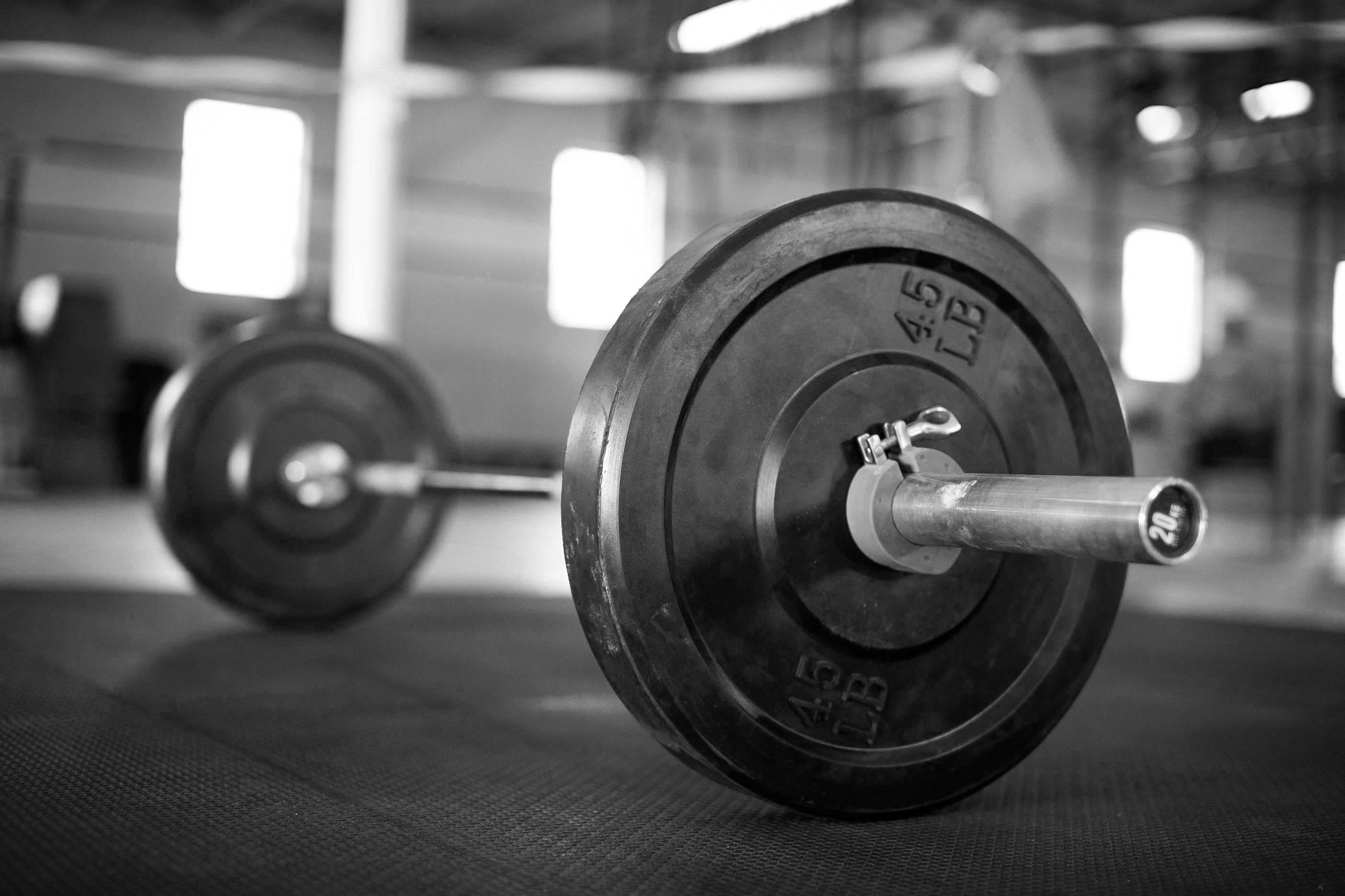 Barbell 桌布 壁紙 — 黑白特寫，槓鈴連槓片放在健身房地板上，背景器材模糊。