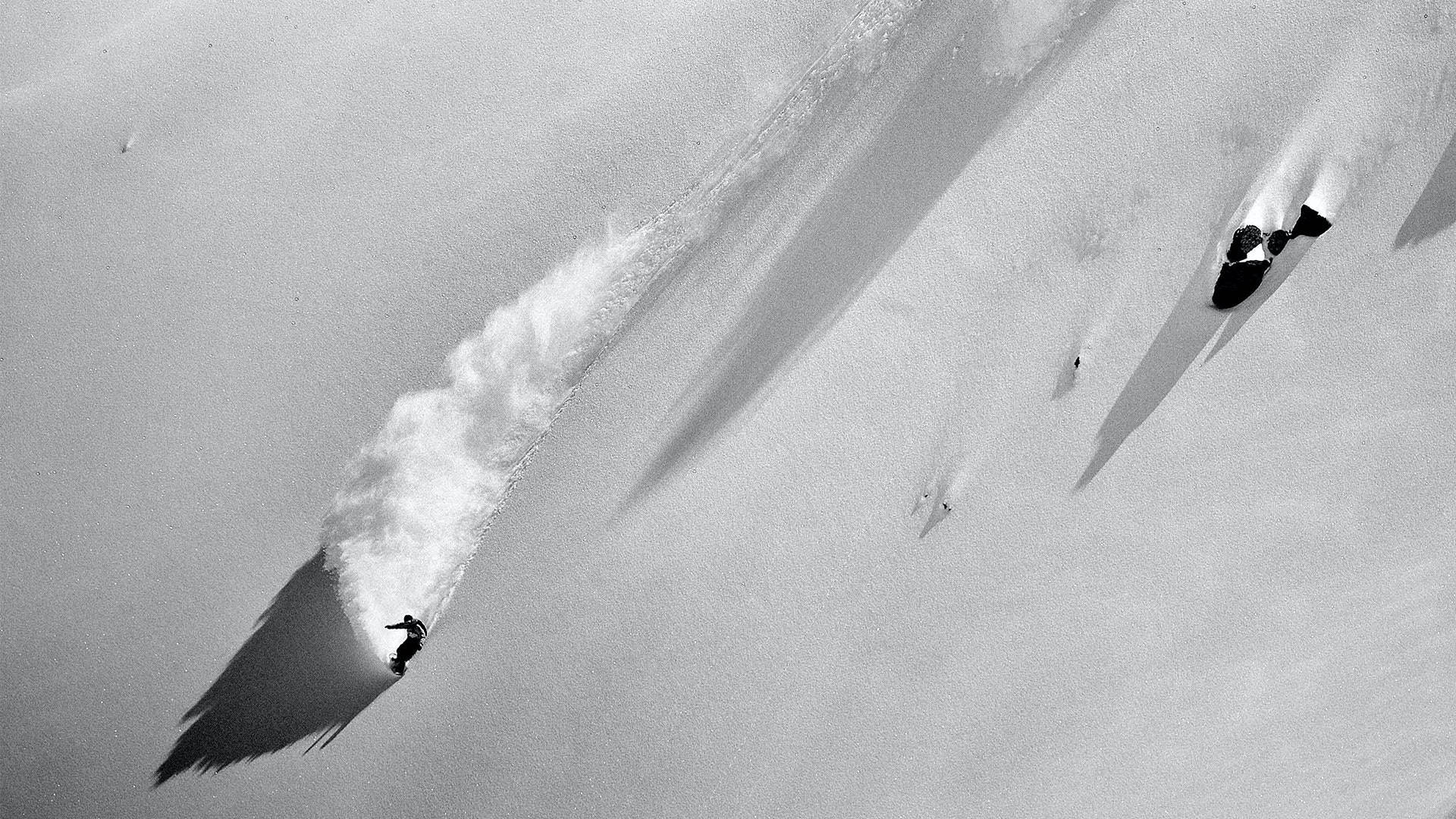 Snowboarding 桌布 壁紙 — 黑白航拍鏡頭，捕捉滑雪板運動員在陡峭的雪坡上雕刻，身後留下粉狀痕跡。
