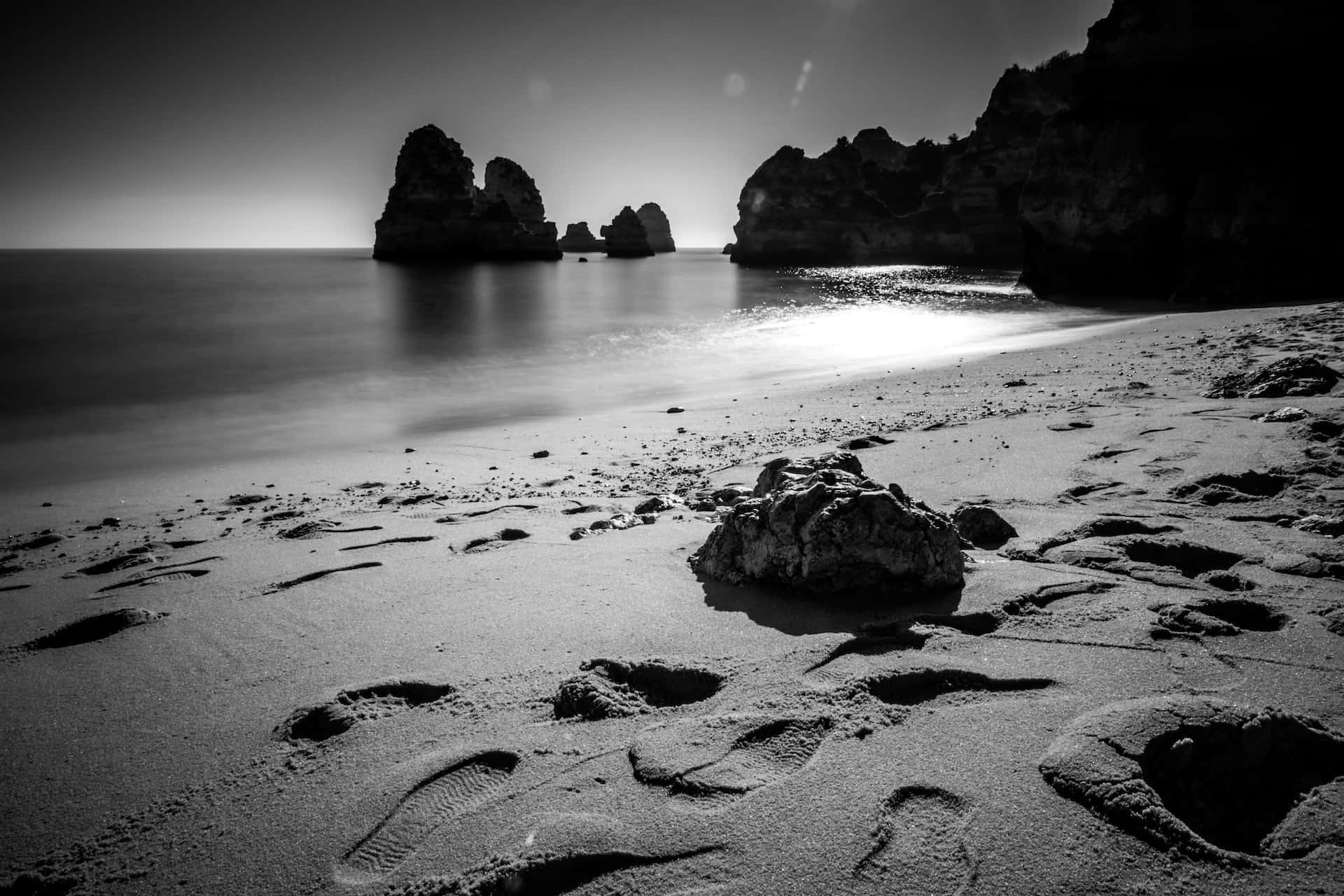 Black And White Beach 桌布 壁紙 — 黑白長曝光海灘場景，沙灘上有腳印，明亮的陽光下戲劇性的岩石 formations。