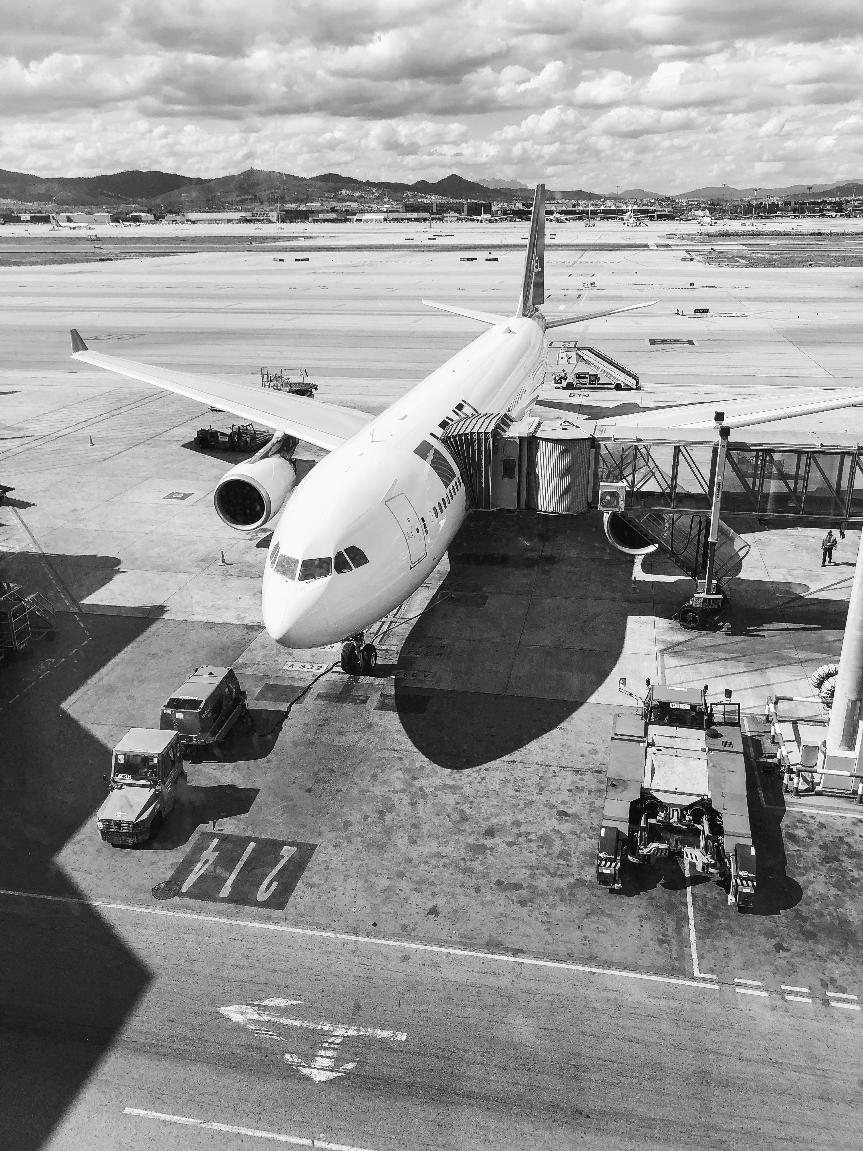 飛機 桌布 壁紙 — 黑白高角度視角，一架大型客機停泊在機場閘口，地面服務車輛和遠山在陰天背景下。
