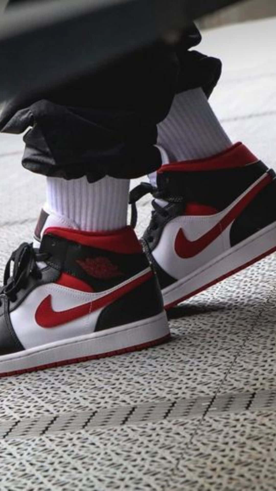 鞋子 桌布 壁紙 — 黑色、紅色和白色Jordan 1運動鞋的特寫，搭配白色襪子和黑色褲子，站在有圖案的人行道上。