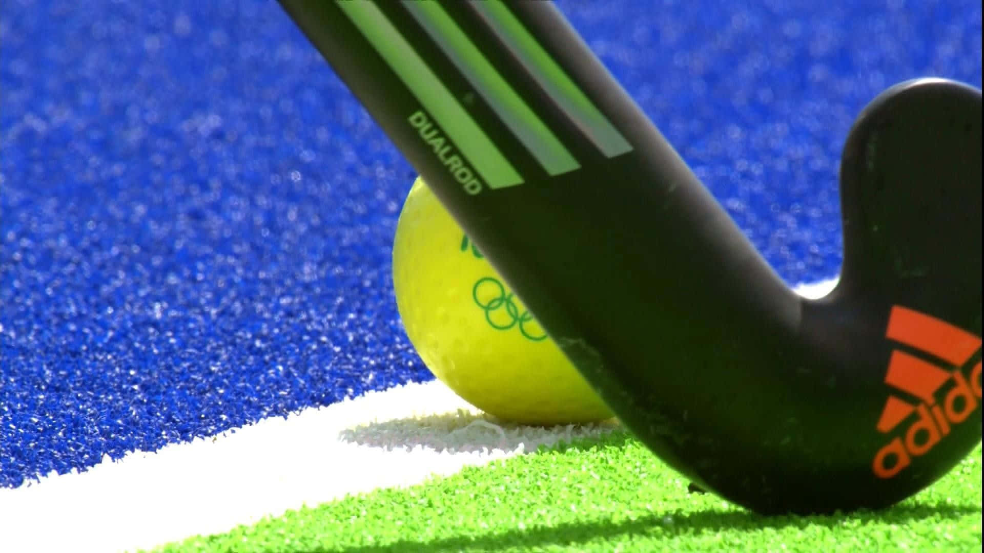 hockey 桌布 壁紙 — 黑色Adidas曲棍球棍的特寫，放在鮮綠色和藍色的人造草地上，附近有一個黃色球。