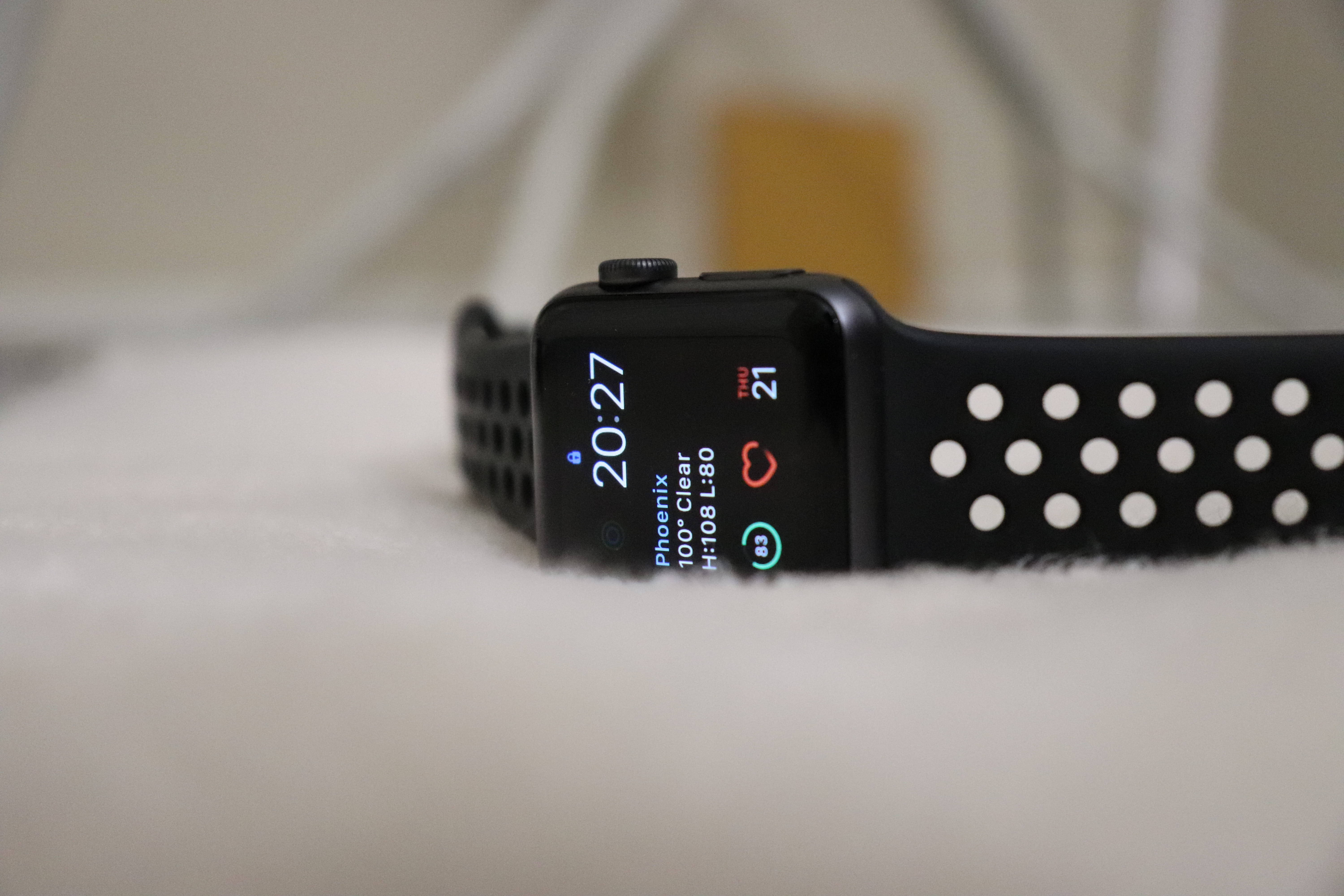 Apple Watch 動態 桌布 壁紙 — 黑色Apple Watch配穿孔錶帶，放置在柔軟的白色表面上，顯示時間、天氣和心率。