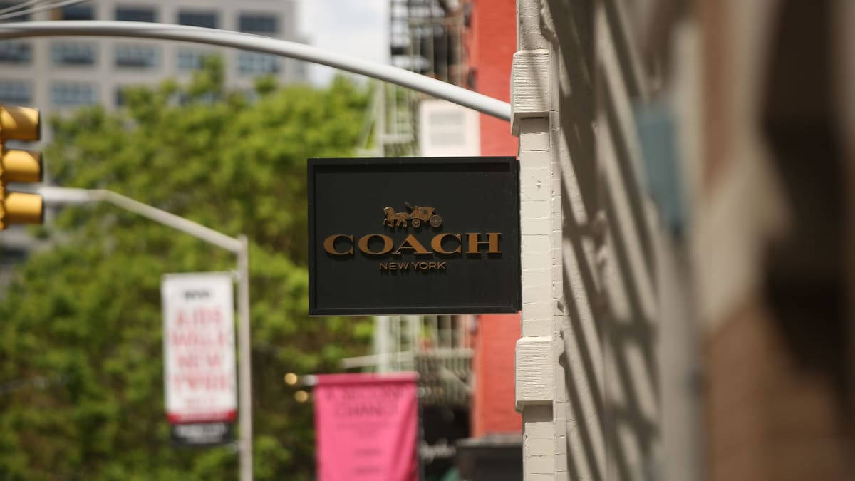 Coach 桌布 壁紙 — 黑色Coach New York商店招牌的特寫，安裝在白色磚牆上，背景是模糊的城市景觀。