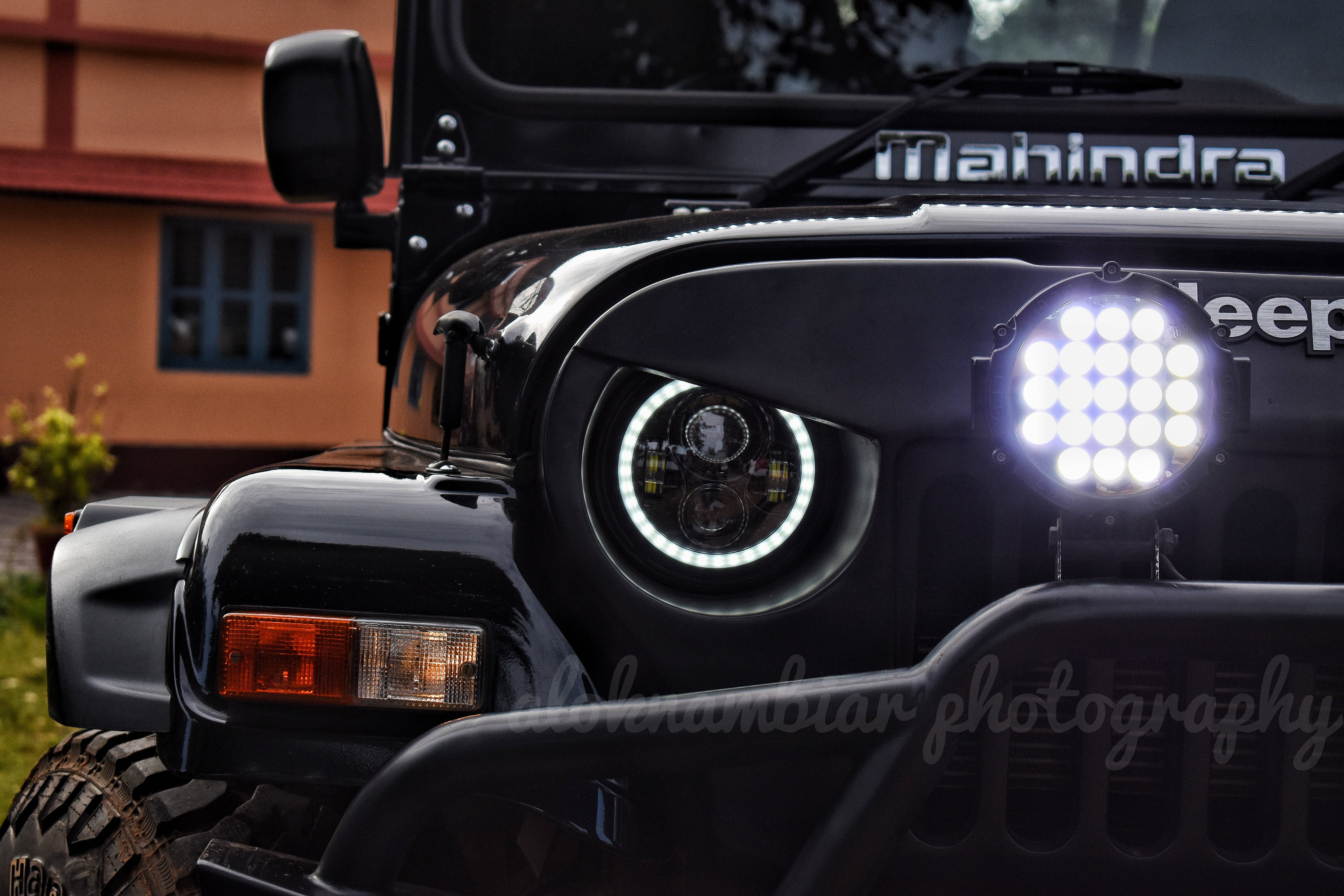 汽車 4K 桌布 壁紙 — 黑色Mahindra Jeep車頭特寫，配備明亮LED頭燈和堅固保險桿，背景為模糊的房屋。