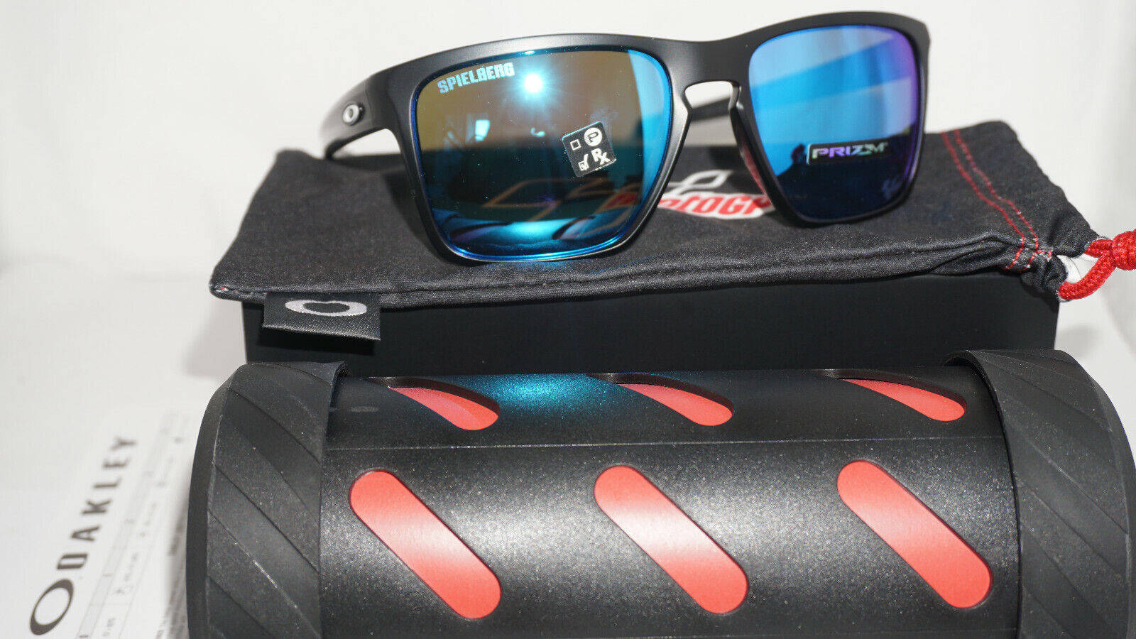 Sunglasses 桌布 壁紙 — 黑色Oakley太陽眼鏡，藍色鏡面鏡片，放在品牌眼鏡盒和紅色點綴的硬殼眼鏡盒上。