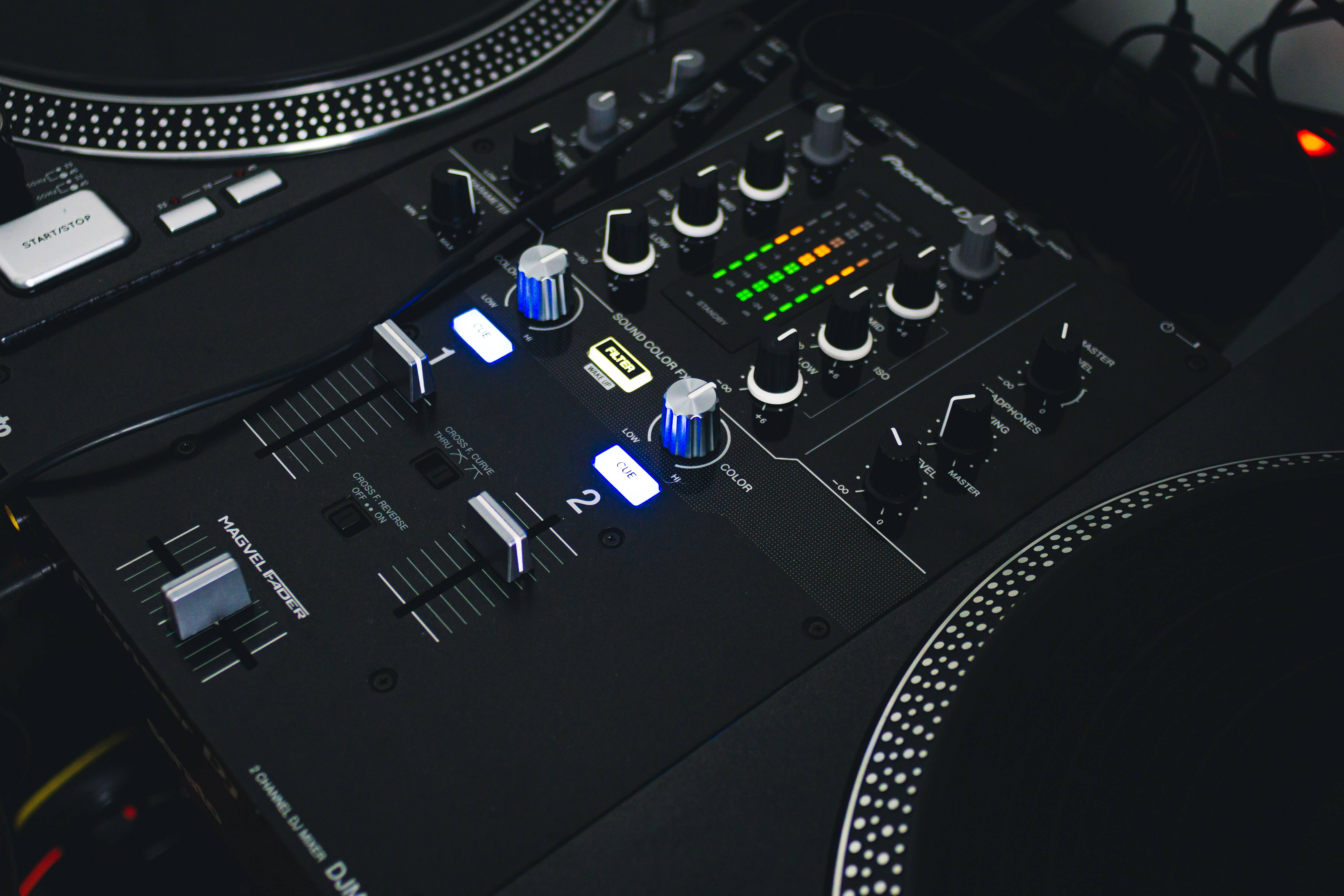 Discord 桌布 壁紙 — 黑色Pioneer DJ混音器特寫，帶有發光藍色旋鈕和綠色LED儀表，位於兩個唱盤之間。